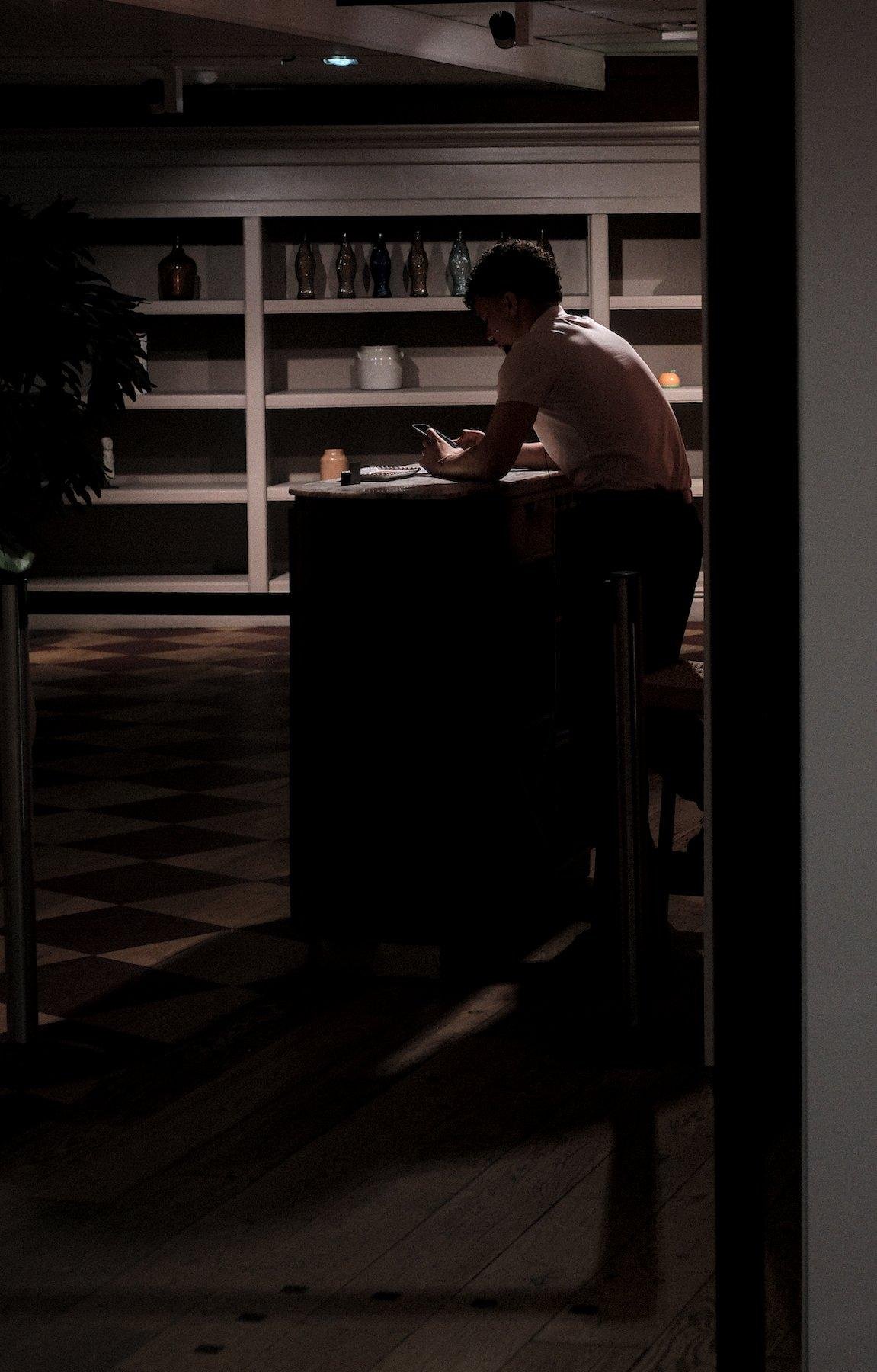 Photo verticale. Dans la pĆ©nombre dāune faible lumiĆØre, un homme est assis sur un tabouret haut Ć un petit comptoir dāaccueil