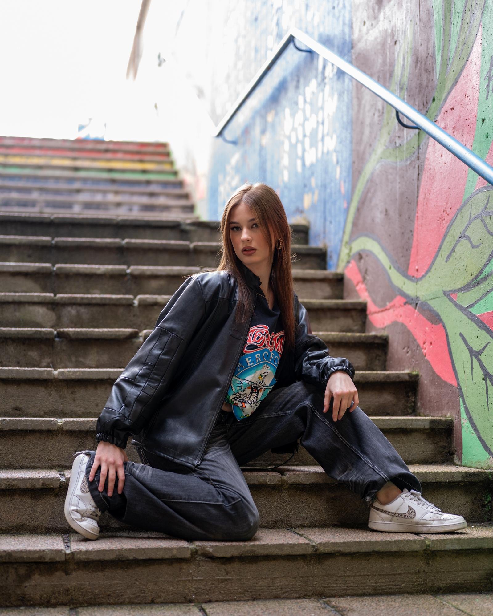 Young woman sitting on stairs wearing a black leather jacket, graphic t-shirt, dark pants, and white sneakers. Background includes a colorful mural on an adjoining wall and rainbow-colored steps.