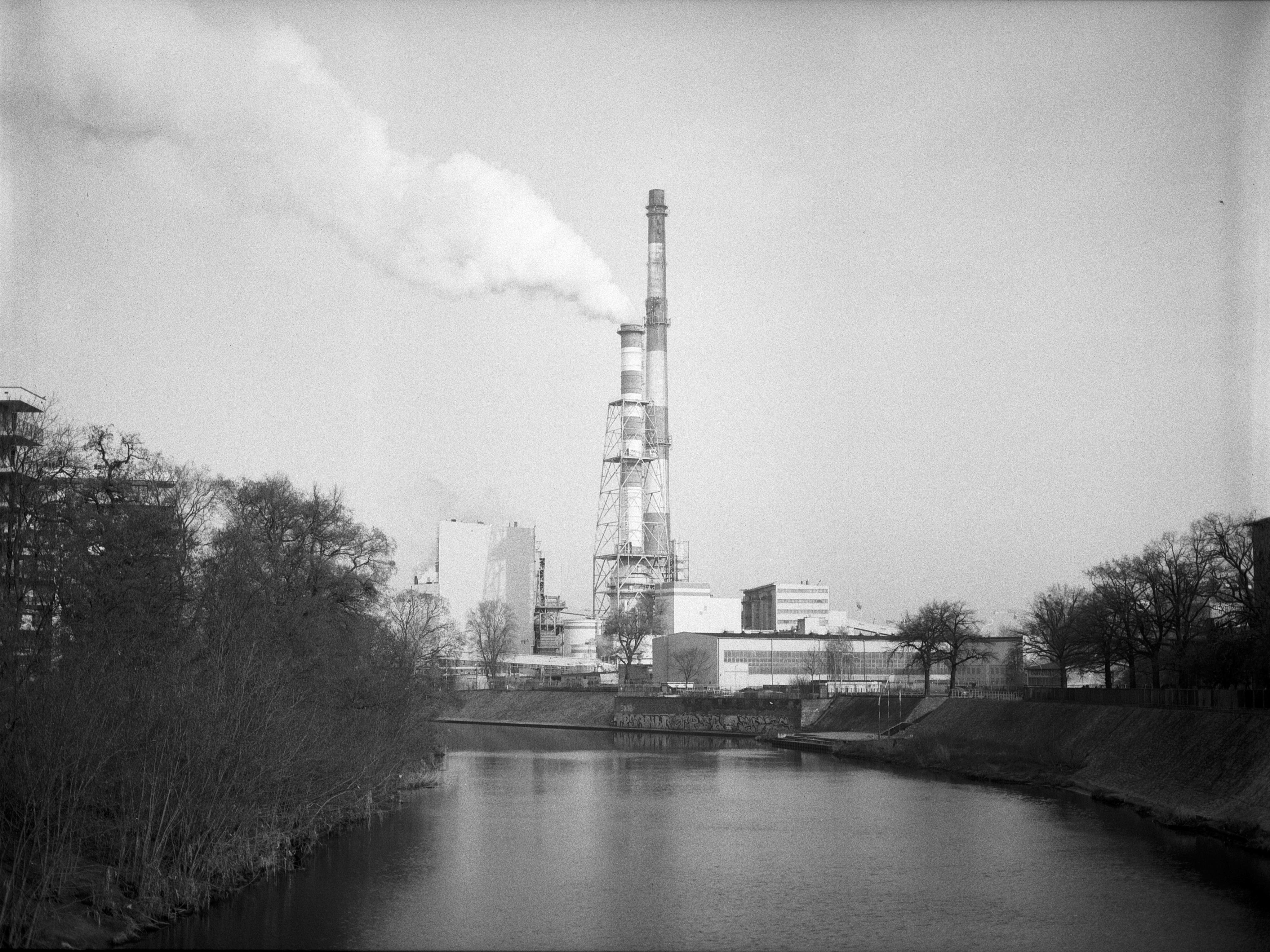 Black and white image of an industrial landscape featuring a large smokestack emitting white smoke in the center. Buildings and storage tanks are visible around the base of the smokestack. A river runs across the foreground, with bare trees lining both sides. The sky is clear, and the overall scene appears stark and industrial.