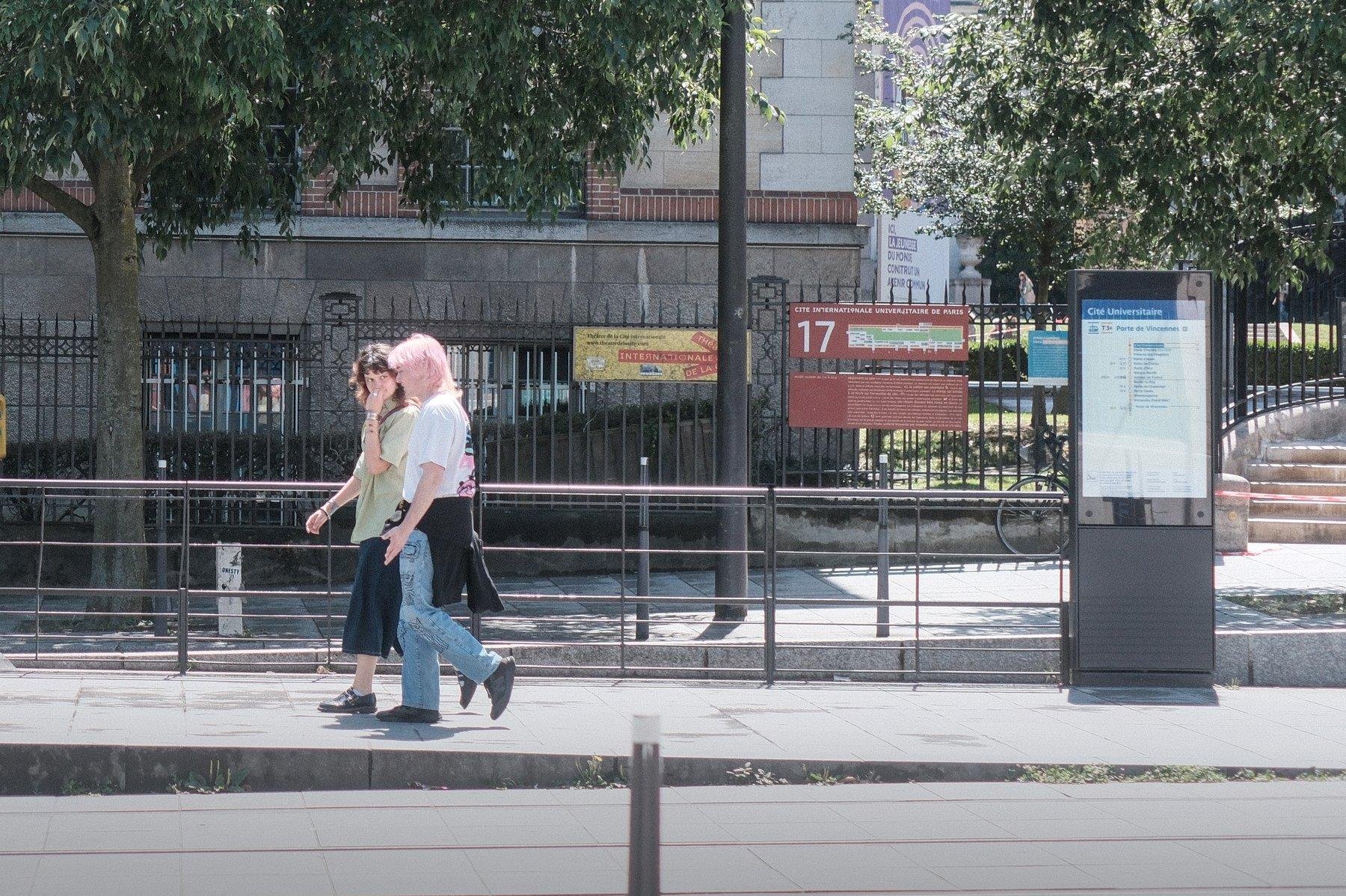 Photo horizontale. 2 jeune femmes marches dans la rue vers la gauche, sous un fort soleil, l’une d’elle a les cheveux rose