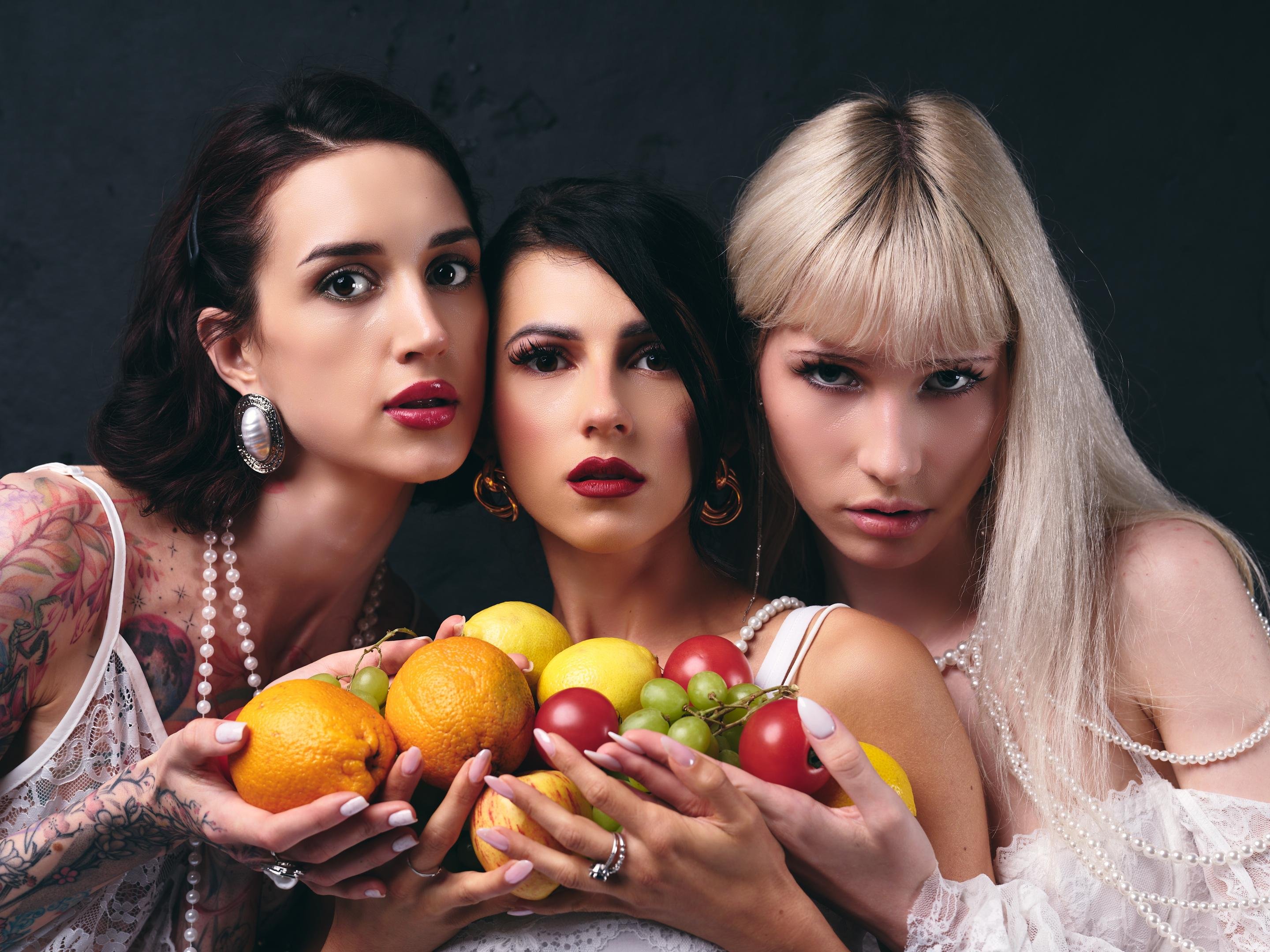 Three women in white lace outfits and pearl jewelry pose closely against a dark background, holding a pile of fruit including oranges, lemons, green grapes, and red plums.