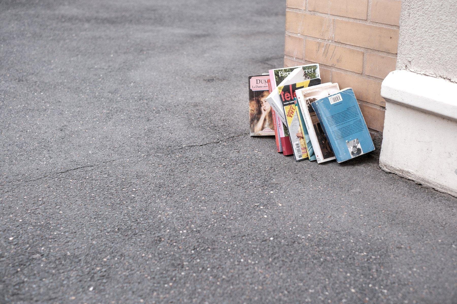 Photo horizontale. Posé sur le coin d’un bâtiment à même le trottoir, des livres de poches en dons