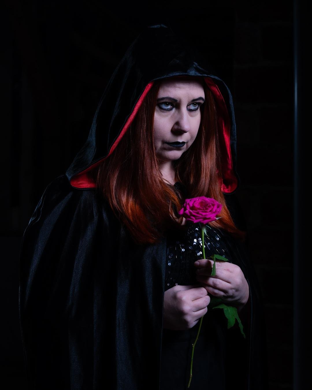 The image shows a person wearing a black cloak with a red lining. They have long reddish hair and are holding a pink rose. The makeup is dark, with black lipstick and eye makeup, giving a gothic or mysterious look. The background is dark, adding to the atmospheric mood.