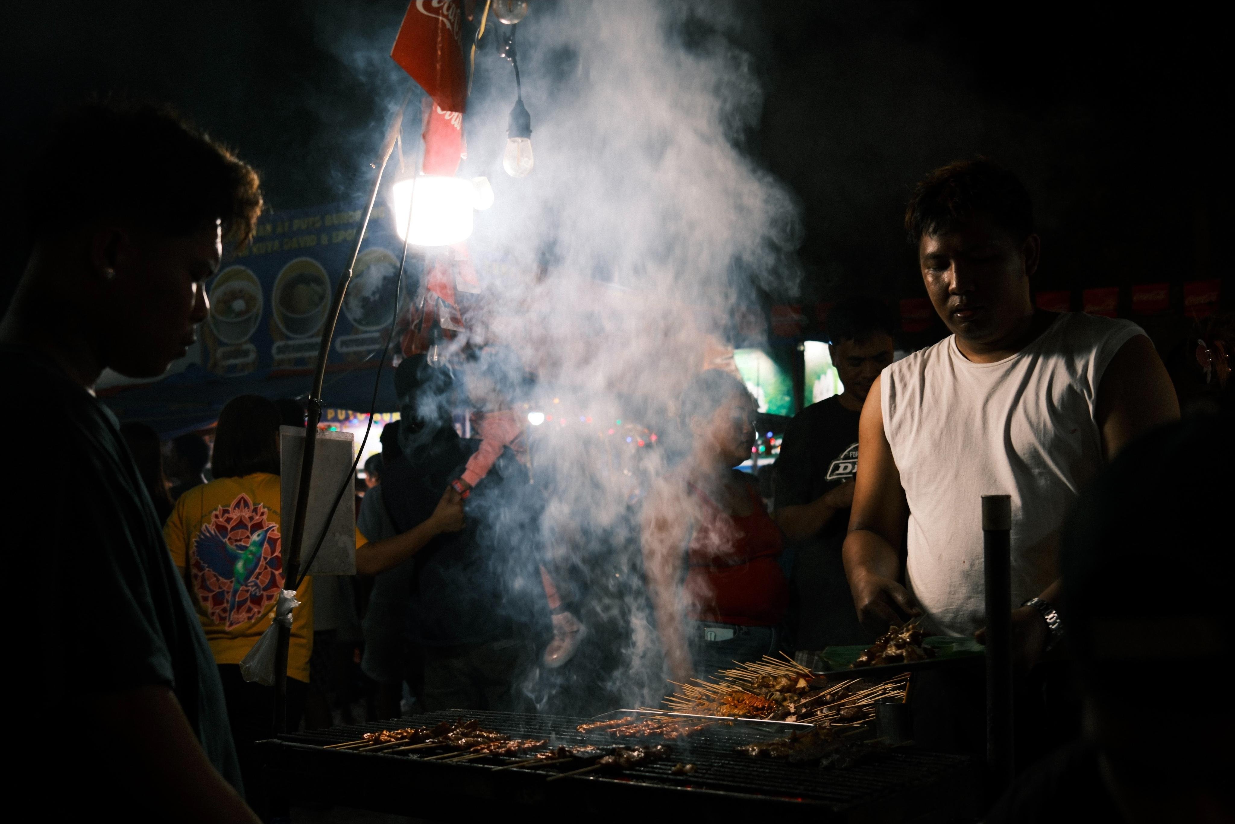 Night street food stall with a charcoal grill in the foreground, skewers of meat cooking as thick smoke rises into a bright overhead light; several people stand around the grill, including a vendor in a white sleeveless shirt on the right, with signs and flags blurred in the background.