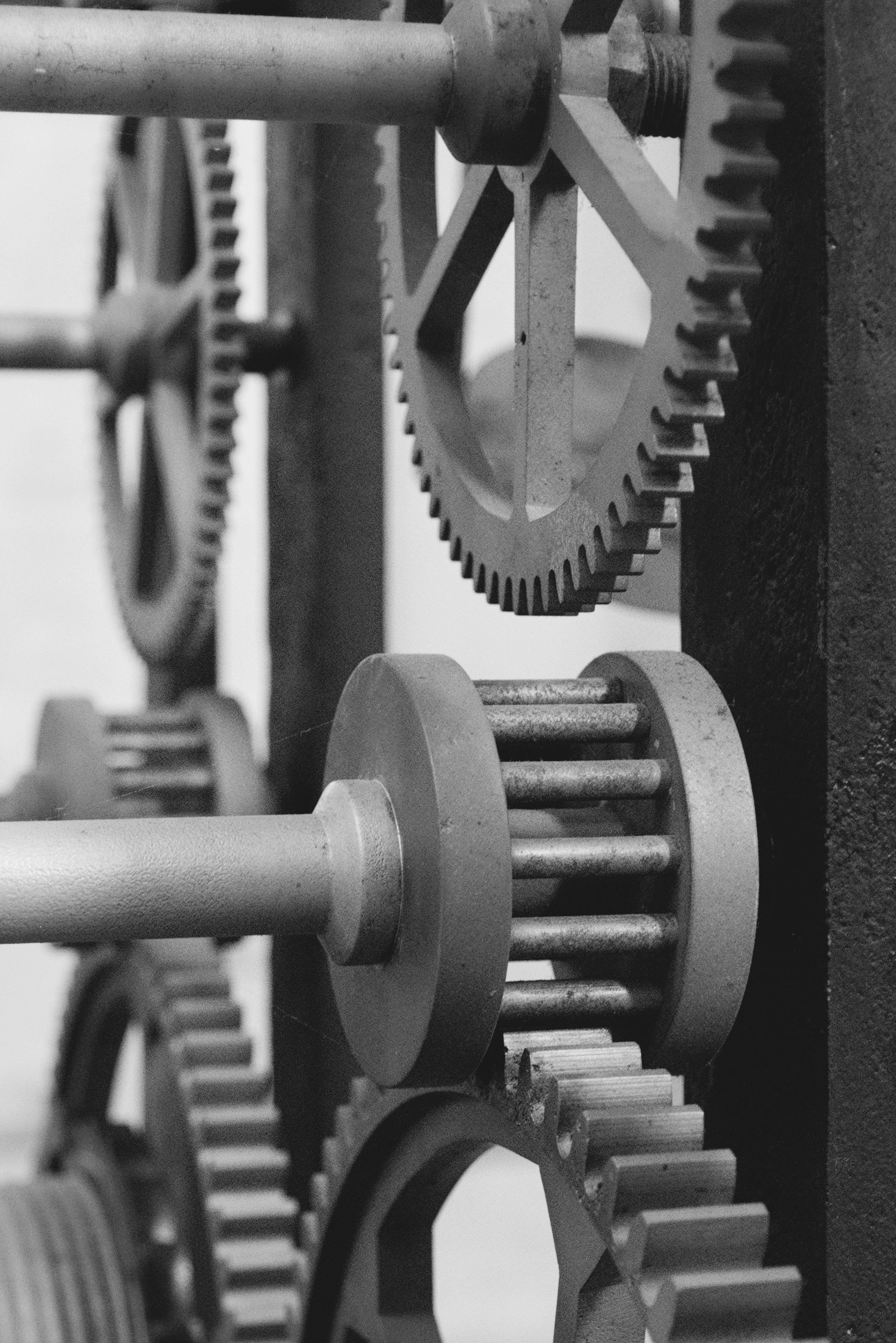 The image shows a close-up of several interlocking gears or cogs, likely part of a mechanical system or machinery. The gears appear to be made of metal and are arranged in a way that suggests they work together to transmit motion or force. The photo is in black and white, emphasizing the texture and details of the gears.