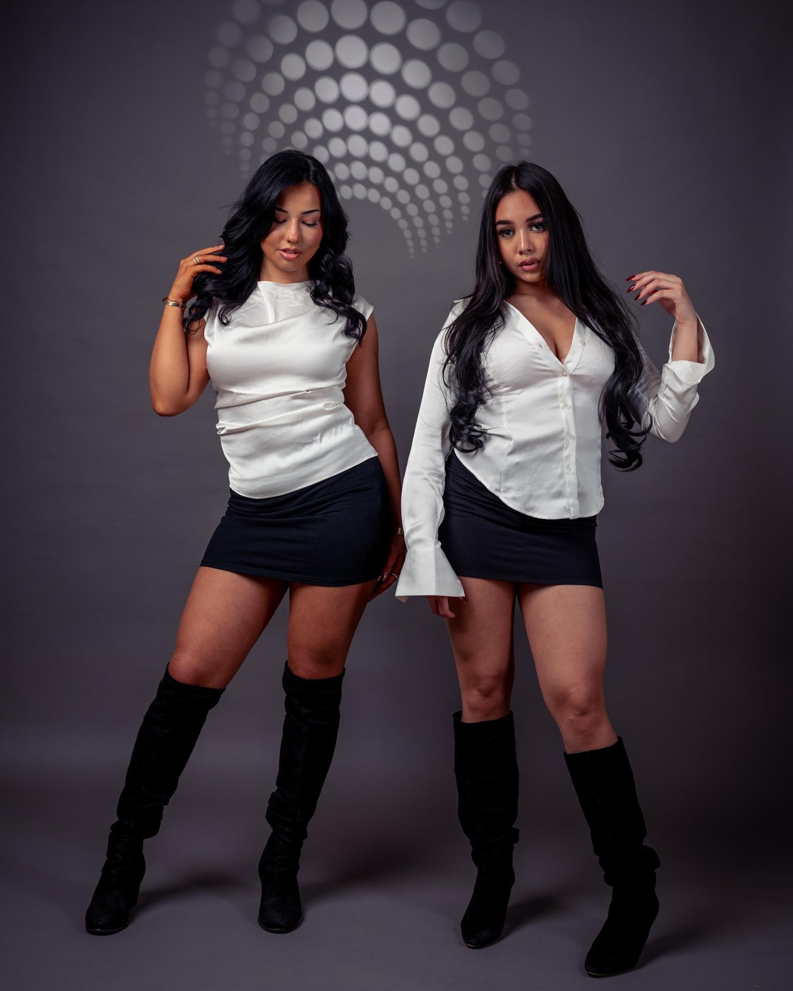 The image shows two women posing in front of a gray background with a pattern of circular lights. They are wearing black skirts, white tops, and black knee-high boots.