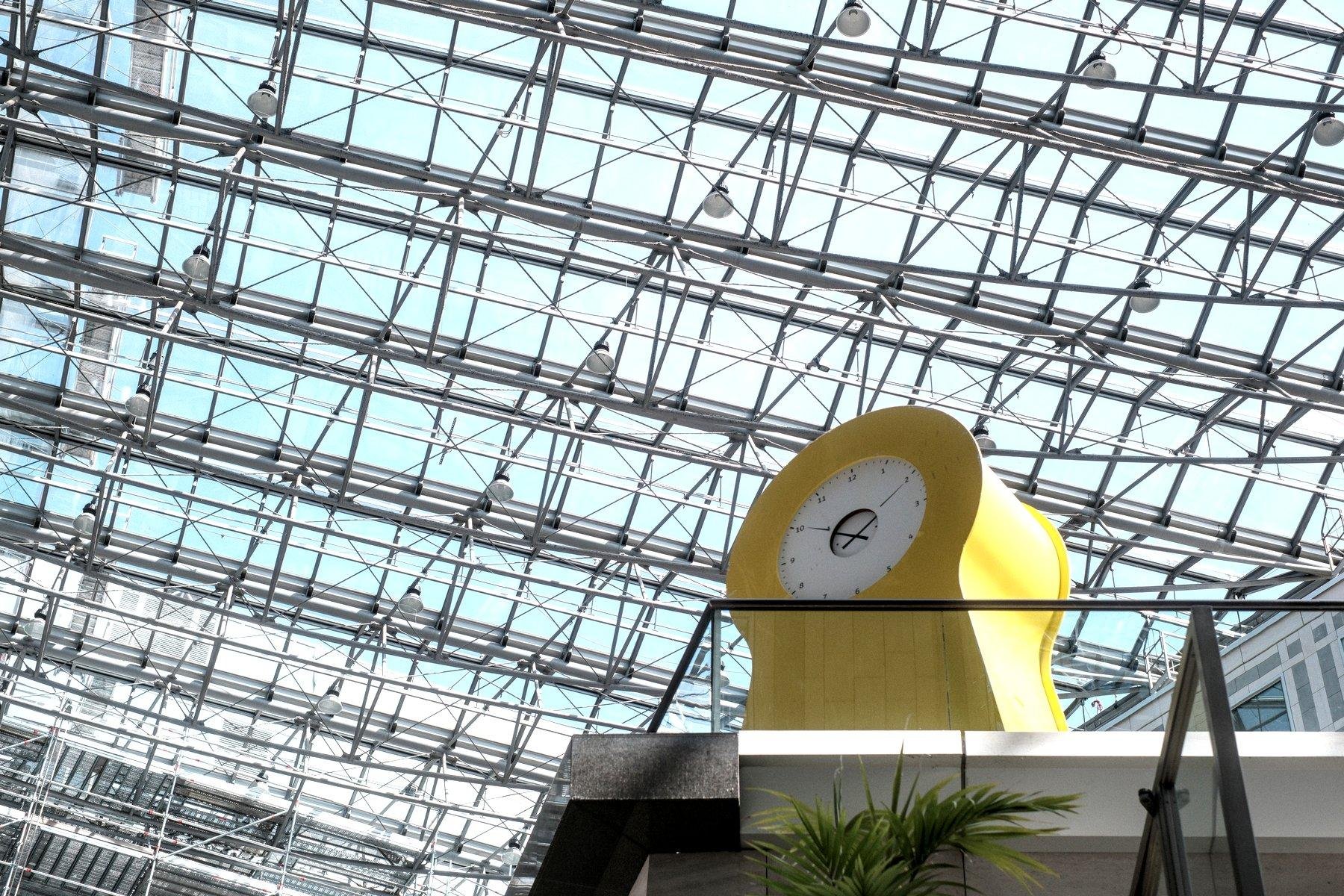 Photo horizontale. Contre plongée sur une horloge jaune au cadran blanc posée sur une mezzanine. Le jaune est celui d’Ikea et le design moderne de l’objet fait penser aussi à Ikea. Encore au dessus, une verrière énorme avec sa structure en métal, laissant passer le bleu du ciel