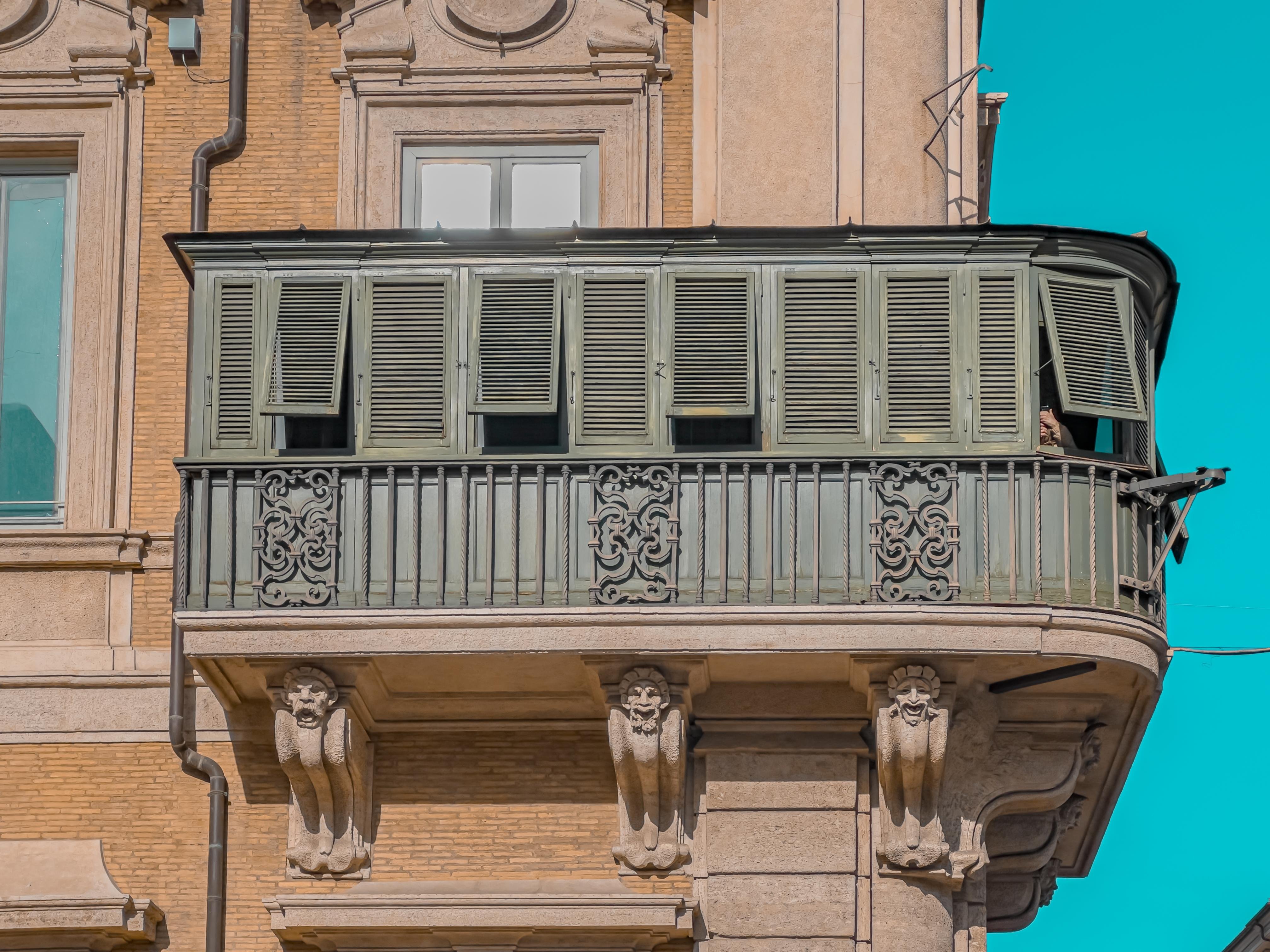Il celebre balcone di Palazzo Bonaparte a Roma, con persiane verdi e raffinati dettagli in ferro battuto, incorniciato da un cielo turchese. Un simbolo di storia e bellezza.