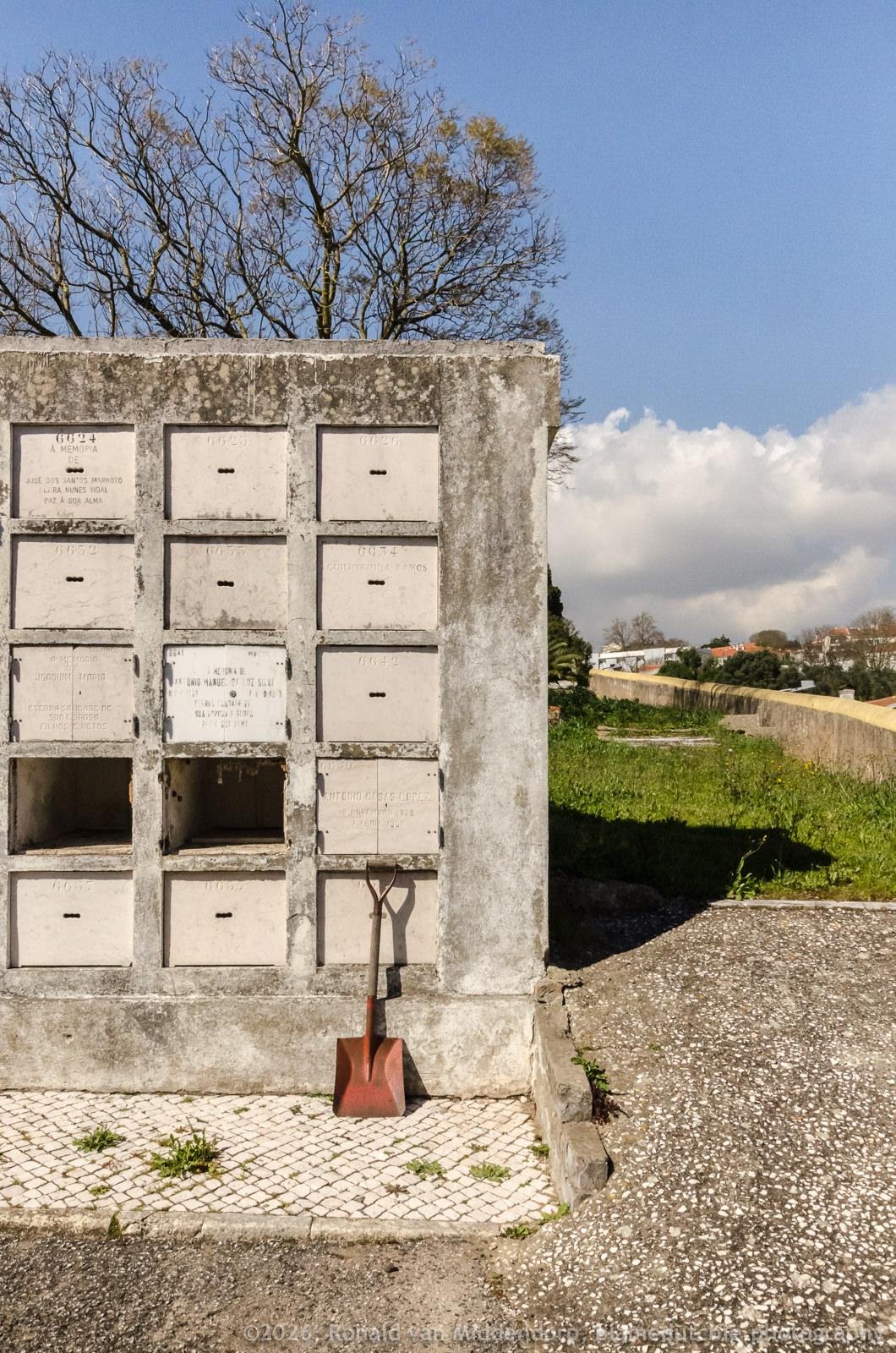 Concrete cemetery mausoleum wall with a grid of sealed niches and engraved plaques; a red shovel leans against the base on a tiled walkway, with leafless trees, grass, and a low stone wall under a blue sky with clouds.