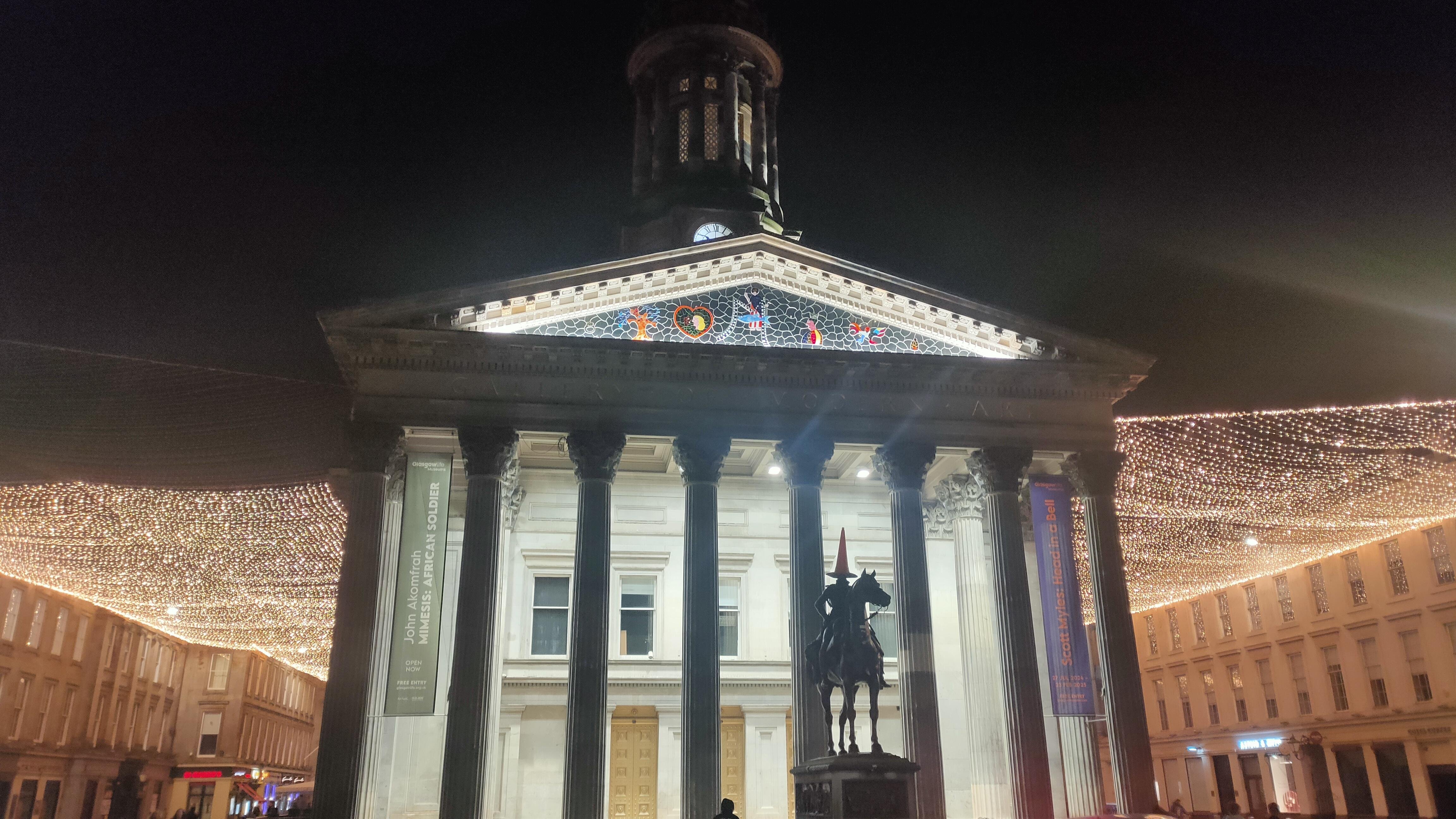 The image shows the Gallery of Modern Art in Glasgow, Scotland, at night. The building is illuminated, showcasing its classical architecture with columns and a tower. In front of the gallery, there is an horse statue, which traditionally has a traffic cone placed on the rider's head as a local humorous tradition. The surrounding area is decorated with festive lights, enhancing the nighttime scene.