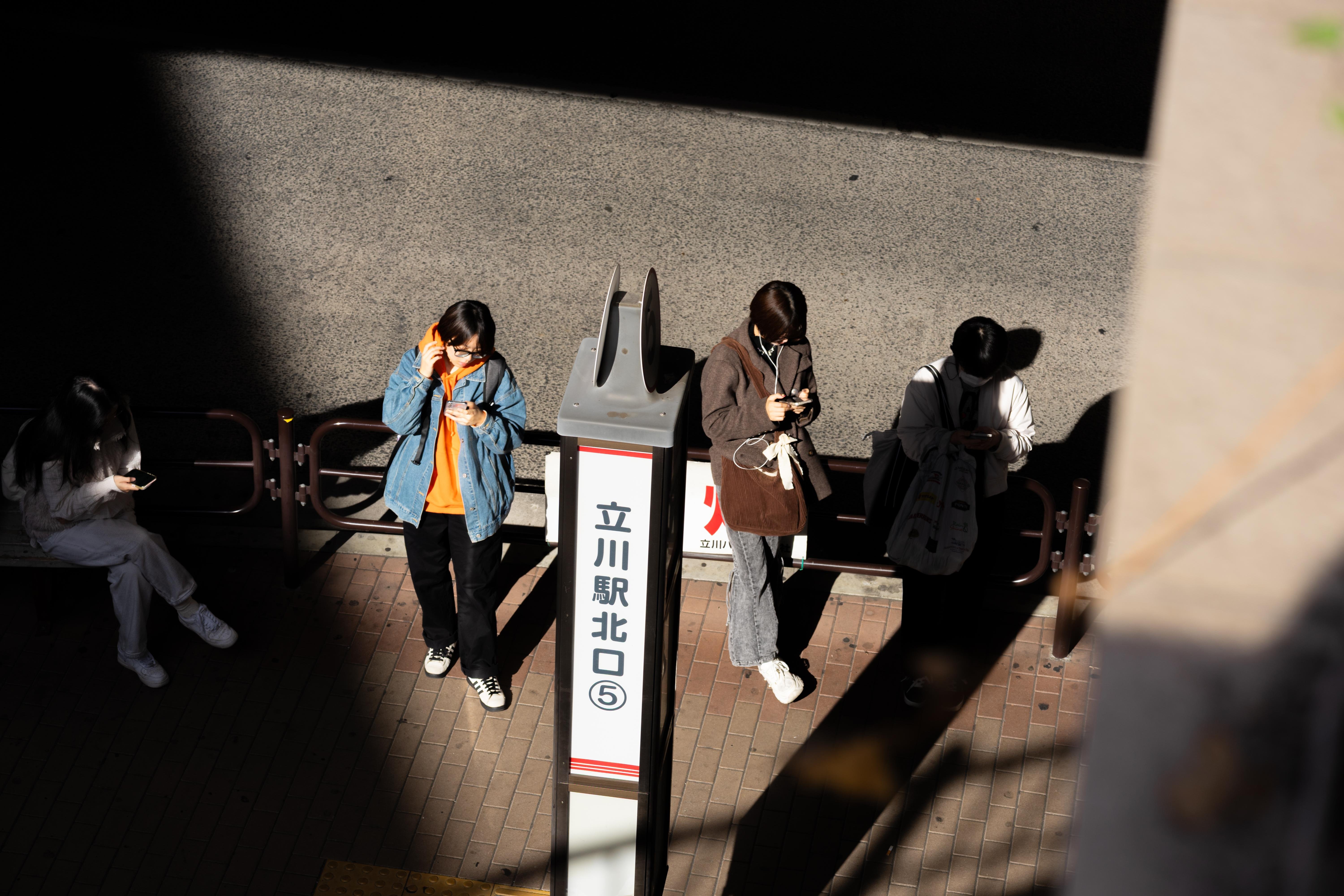 Four people stand on a sunlit sidewalk next to a sign with Japanese text. Each person is engrossed in their phone, wearing casual clothing. Shadows create contrasting light and dark areas. The scene suggests a public waiting area.