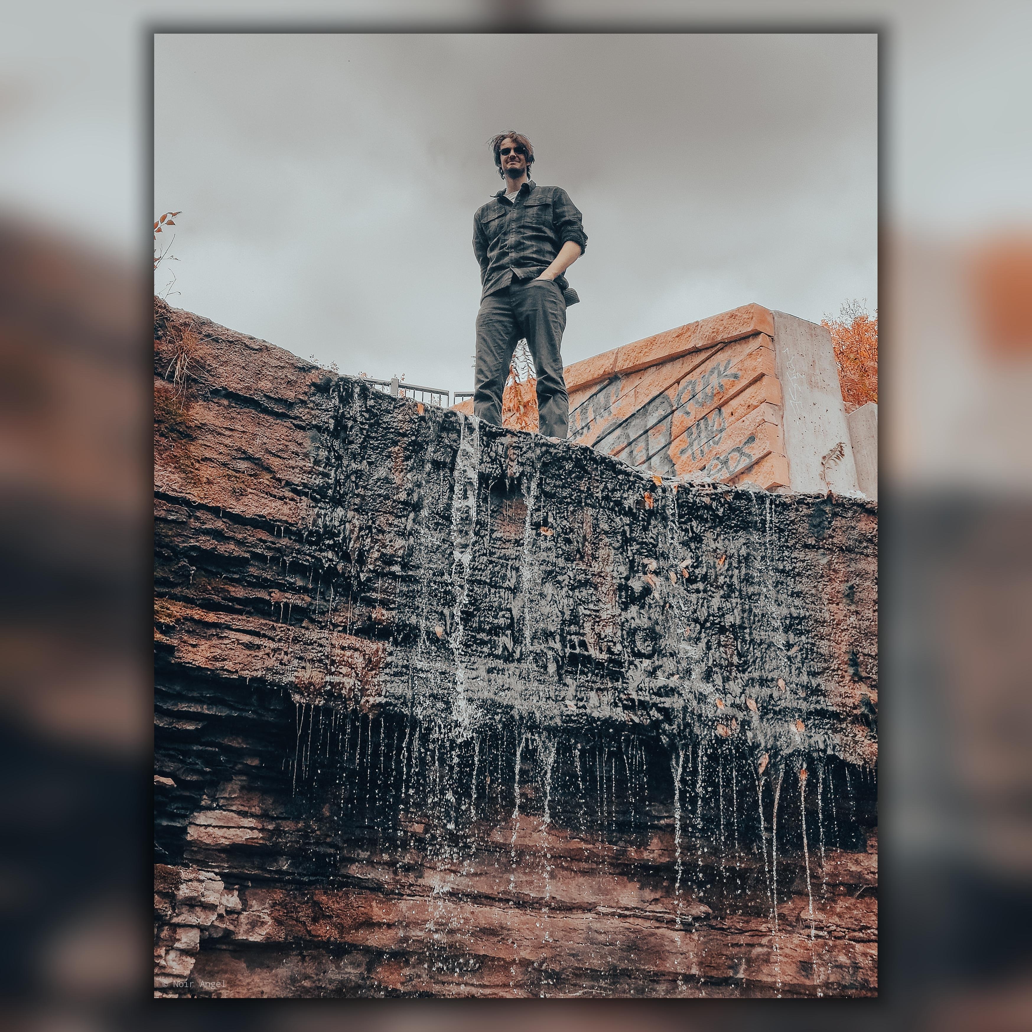 Man standing on a rock ledge with water streaming down its face. Overcast sky above, with graffiti visible on concrete structures in the background. Wearing a dark plaid shirt and pants. Brown and gray tones dominate the scene.