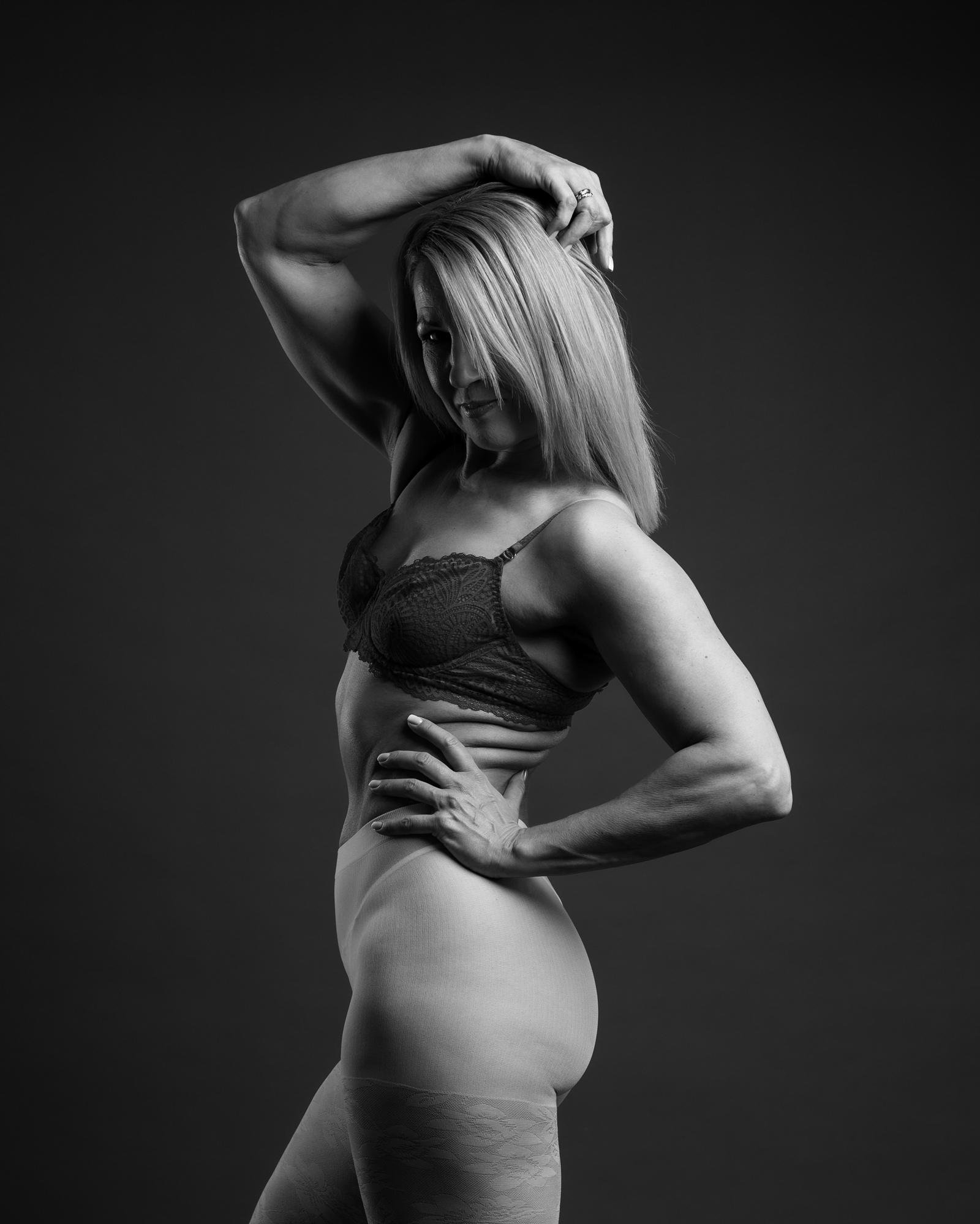 The image is a black and white portrait of a woman posing in a studio setting. She has her hand on her hip and one arm raised above her head, showing a confident stance. She is wearing a bra and sheer stockings, and her hair falls to one side of her face. The mood of the image is artistic and expressive.
