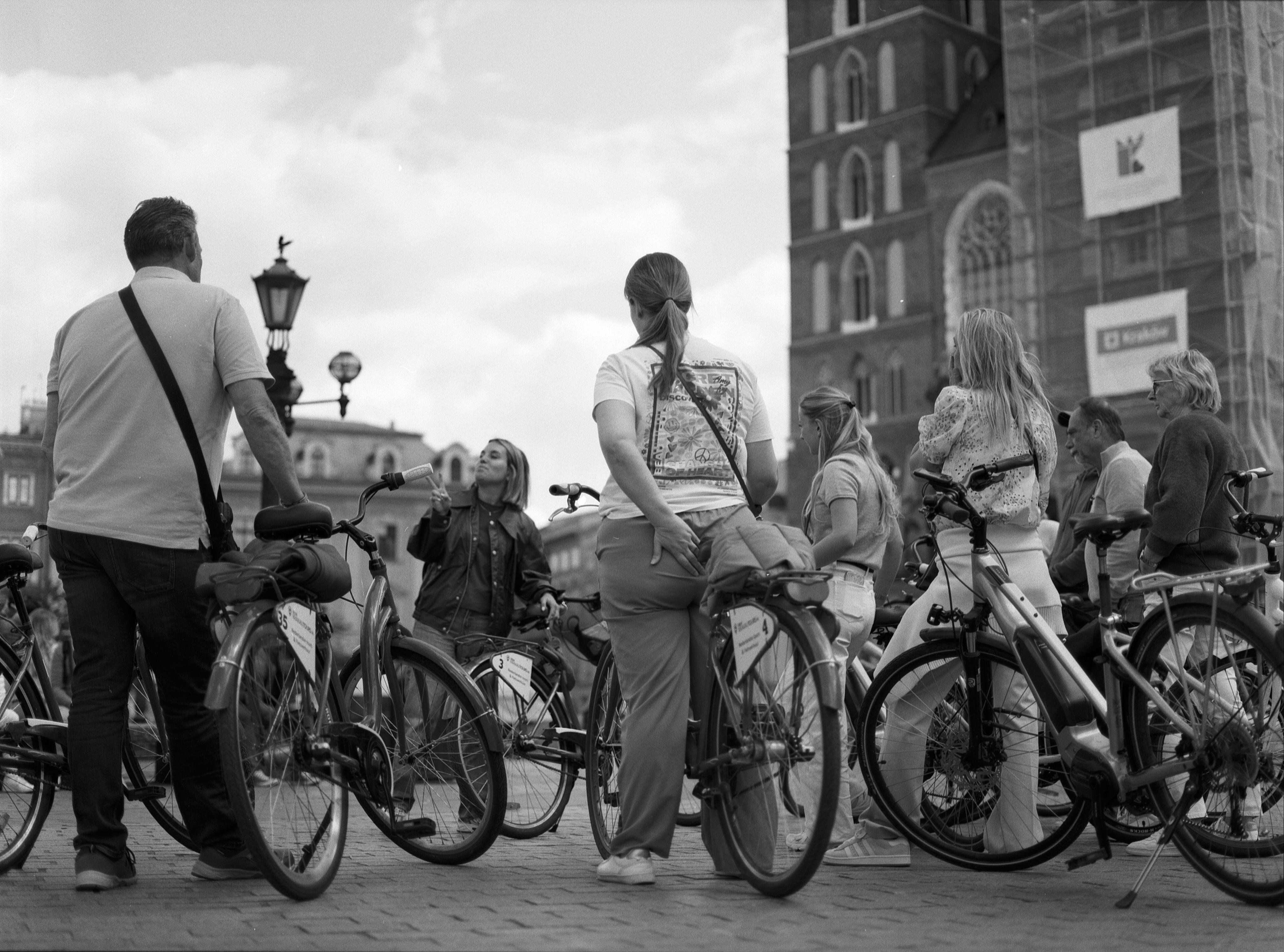 People on bicycles gathered in a city square, facing an individual speaking and gesturing. An old building with scaffold and advertisements looms in the background. The sky is cloudy, and a lamppost and historical architecture are visible. Black and white photograph.