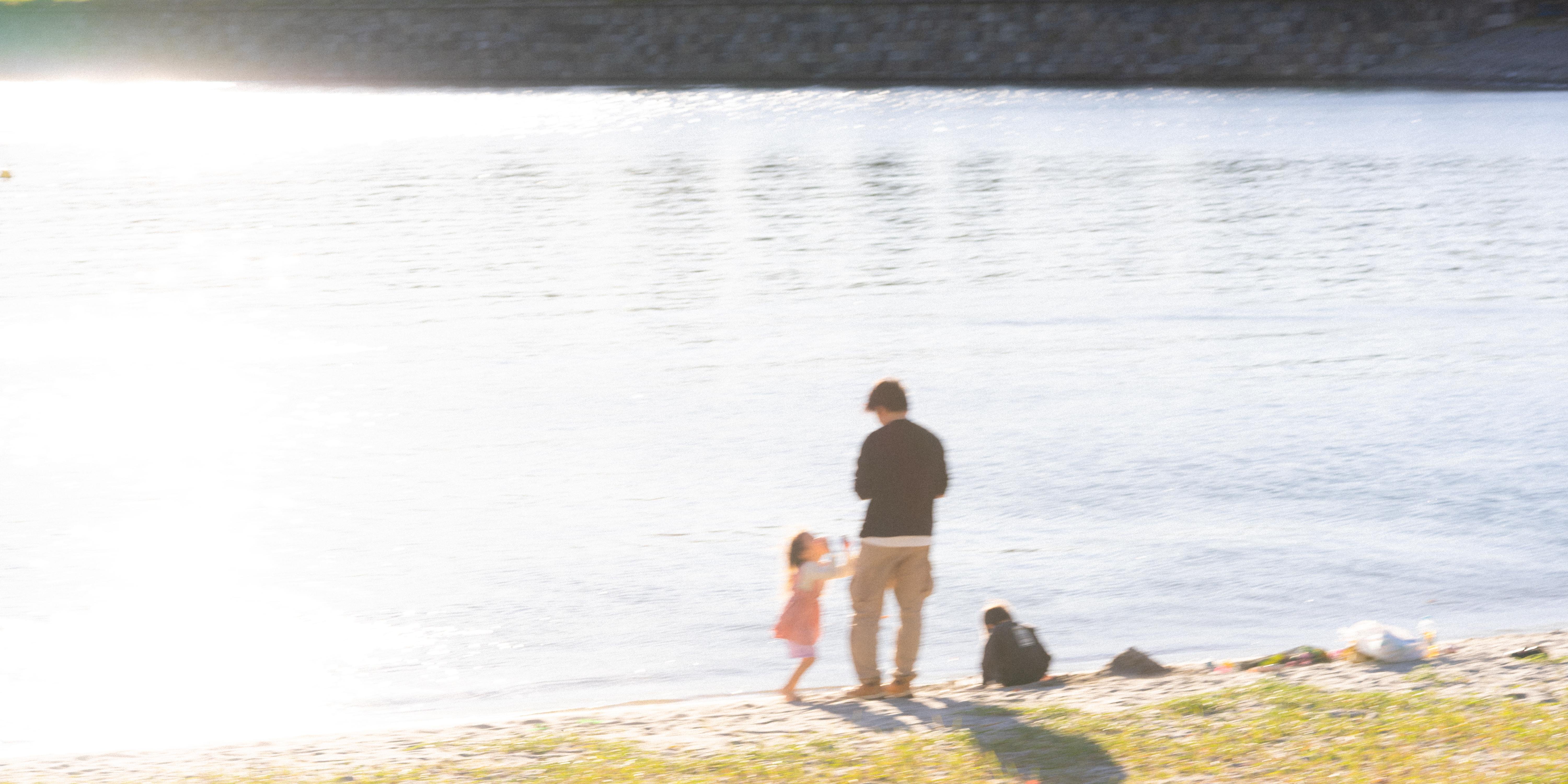 A man and a child stand on a sunlit beach by a calm body of water. The child, wearing a pink dress, reaches towards the man, who is dressed in a black shirt and tan pants. A small black dog sits nearby, while bags and items lie on the sand. The background features a stone wall lining the water.