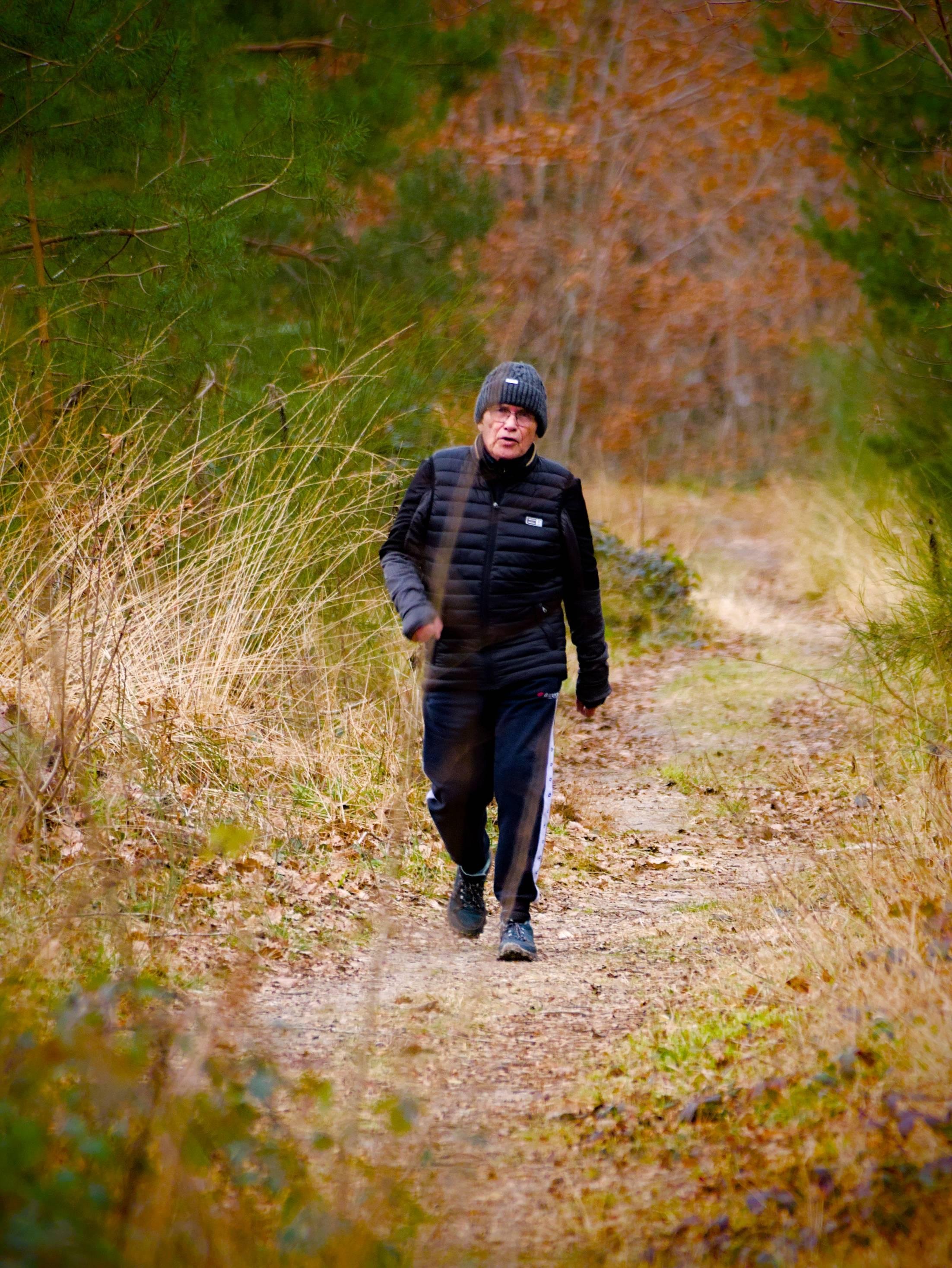 L'image montre une personne marchant sur un sentier dans un environnement naturel. La personne porte une veste noire, un pantalon noir et un bonnet. Le sentier est entouré d'herbe et d'arbres, ce qui suggère qu'il s'agit d'une zone boisée.