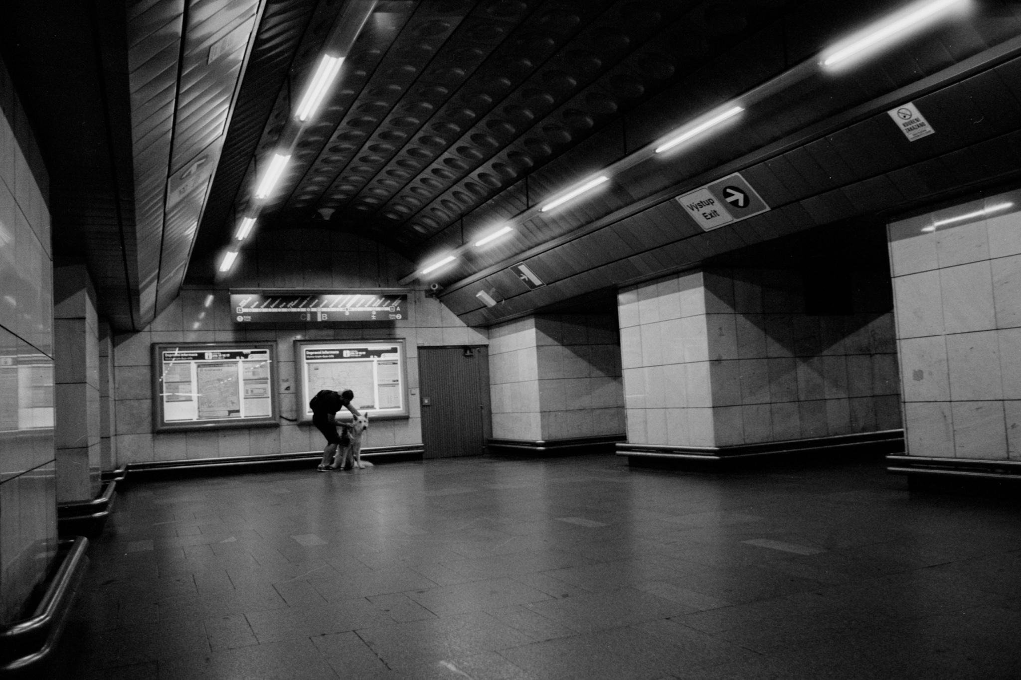 Black-and-white photo of a mostly empty underground transit station with tiled walls, a curved ceiling with fluorescent lights, wall-mounted information boards, and a lone adult bending toward a small child near the boards; directional arrow signs hang overhead.