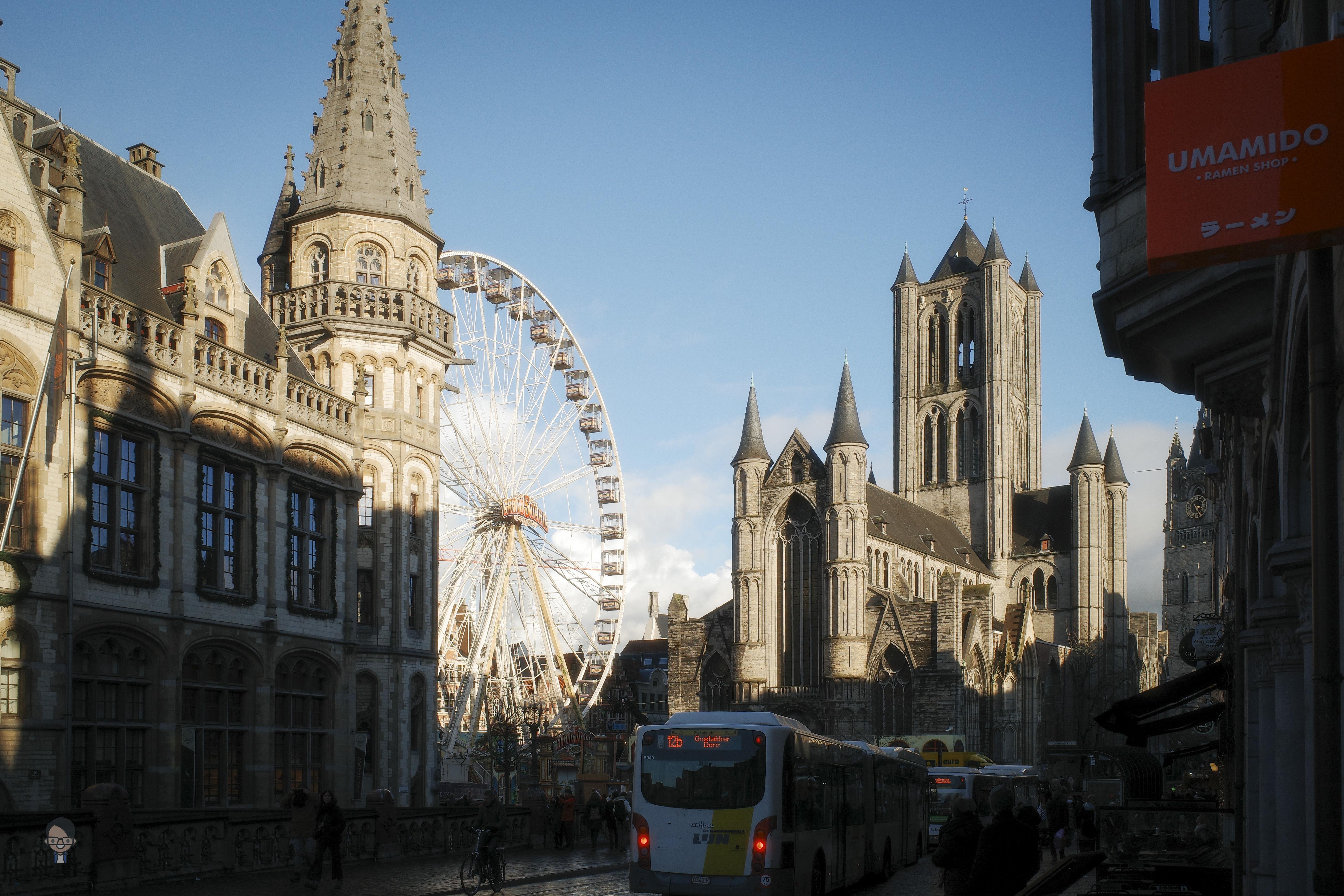The image depicts a cityscape with notable architectural elements. There is a Ferris wheel visible in the background, along with prominent Gothic-style buildings, such as a cathedral with pointed spires. A bus is seen on the street, and there are people walking nearby. Additionally, there's a sign for "UMAMIDO" on the right.