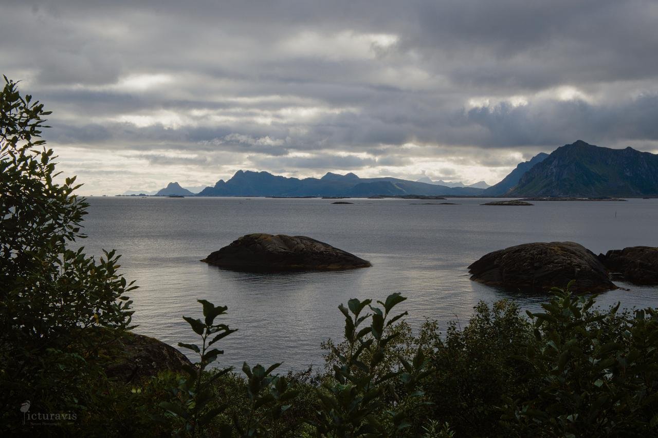 A moody photo of a rugged island landscape with a cloudy sky. Dark green shrubs are in the front, the grey sea with a few grey rocks near the coast in the middle, and a low mountainscape on the horizon. The sun shines through less dense parts of the cloud.