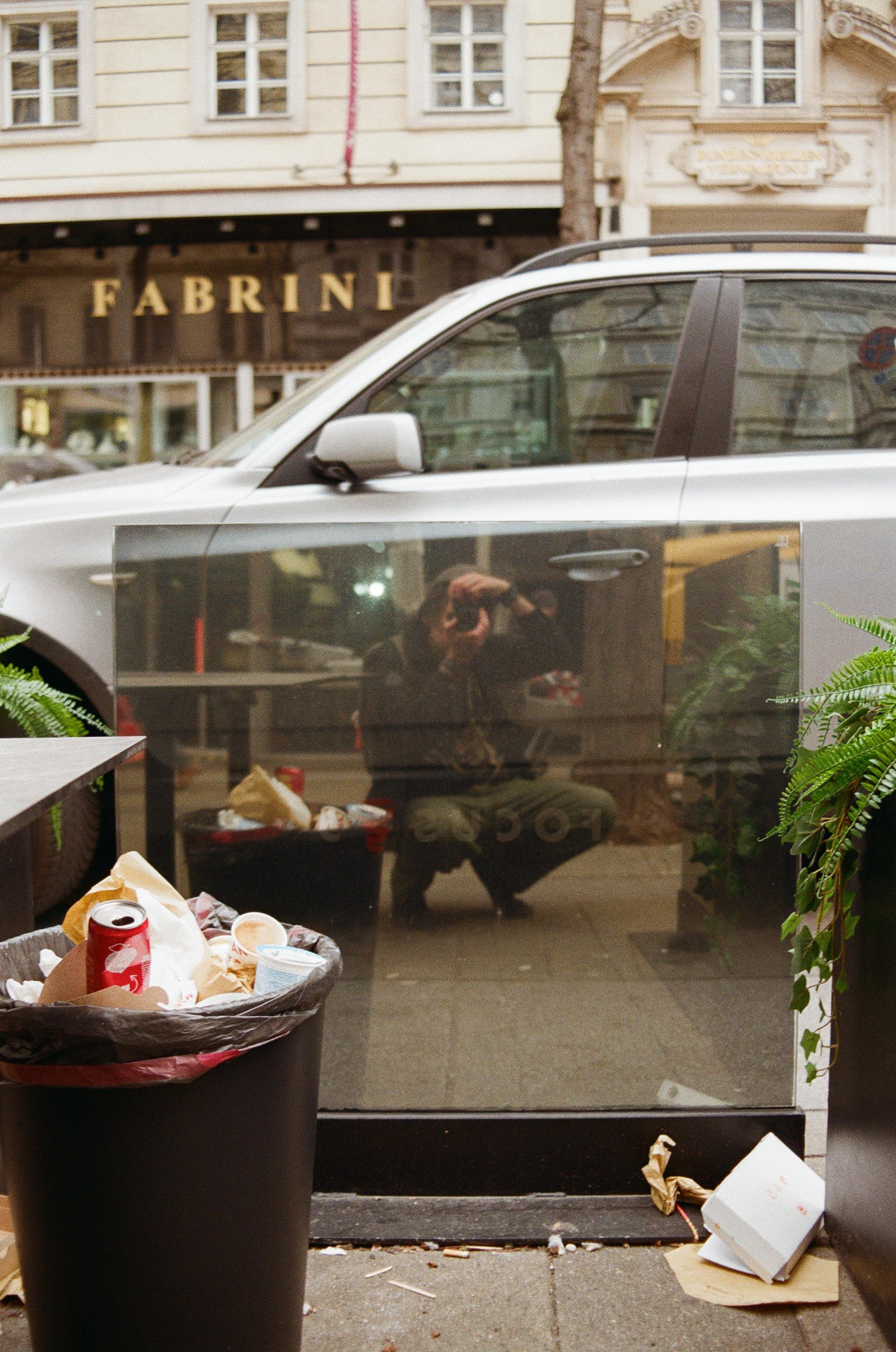 The image shows a reflection of a person holding a camera, taking a picture. There's an overflowing trash can with various items like drink cans and paper. Nearby, there's a silver car parked with a reflection on the side. In the background, there's a building with the word "FABRINI" visible on it. The scene appears to be in an urban setting with elements of litter and plants around.