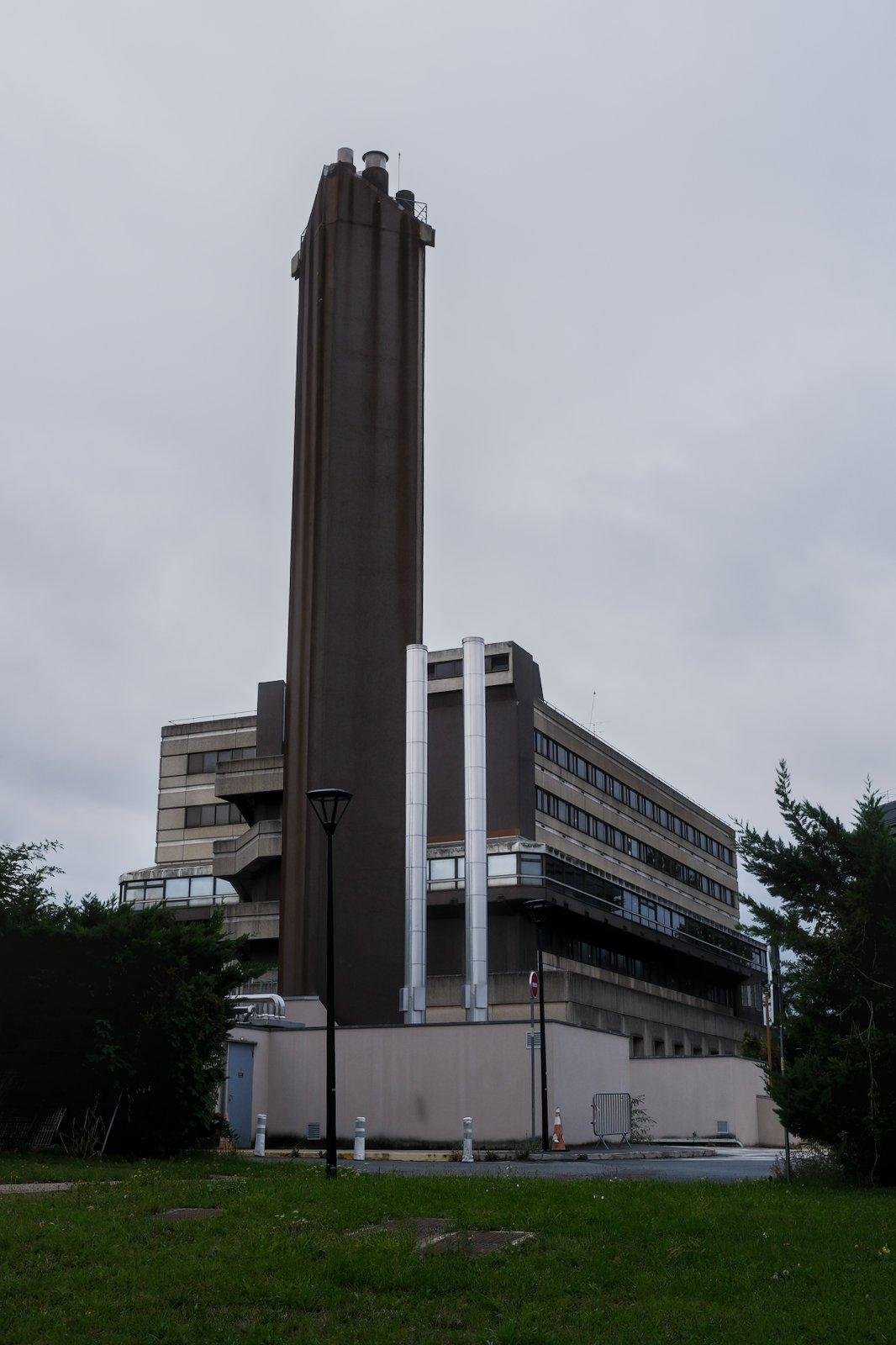 Photo format portrait montrant un bâtiment industriel de couleur beige sale, avec devant, une énorme tour marron très foncé regroupant un ensemble de cheminées. À droit de la tour, 2 autres tuyaux chromés verticaux. À gauche, on voit des escaliers extérieurs. De la pelouse au premier plan, le tout sous un ciel gris