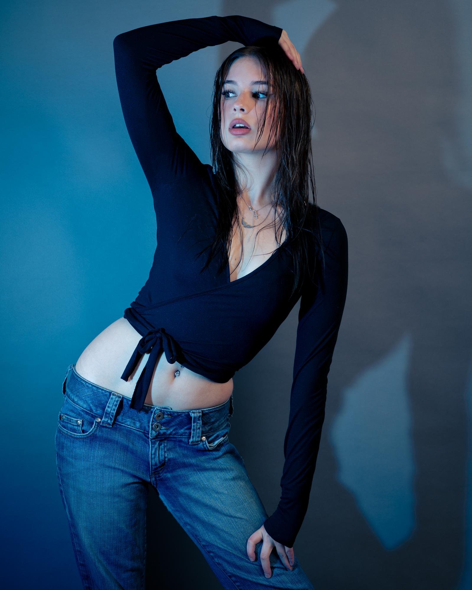 Woman with wet hair posing against a blue background, wearing a black long-sleeve wrap top and blue jeans. She has her left hand raised touching the side of her head while her right hand rests on her hip. The lighting creates a shadow on the background, adding depth to the image.