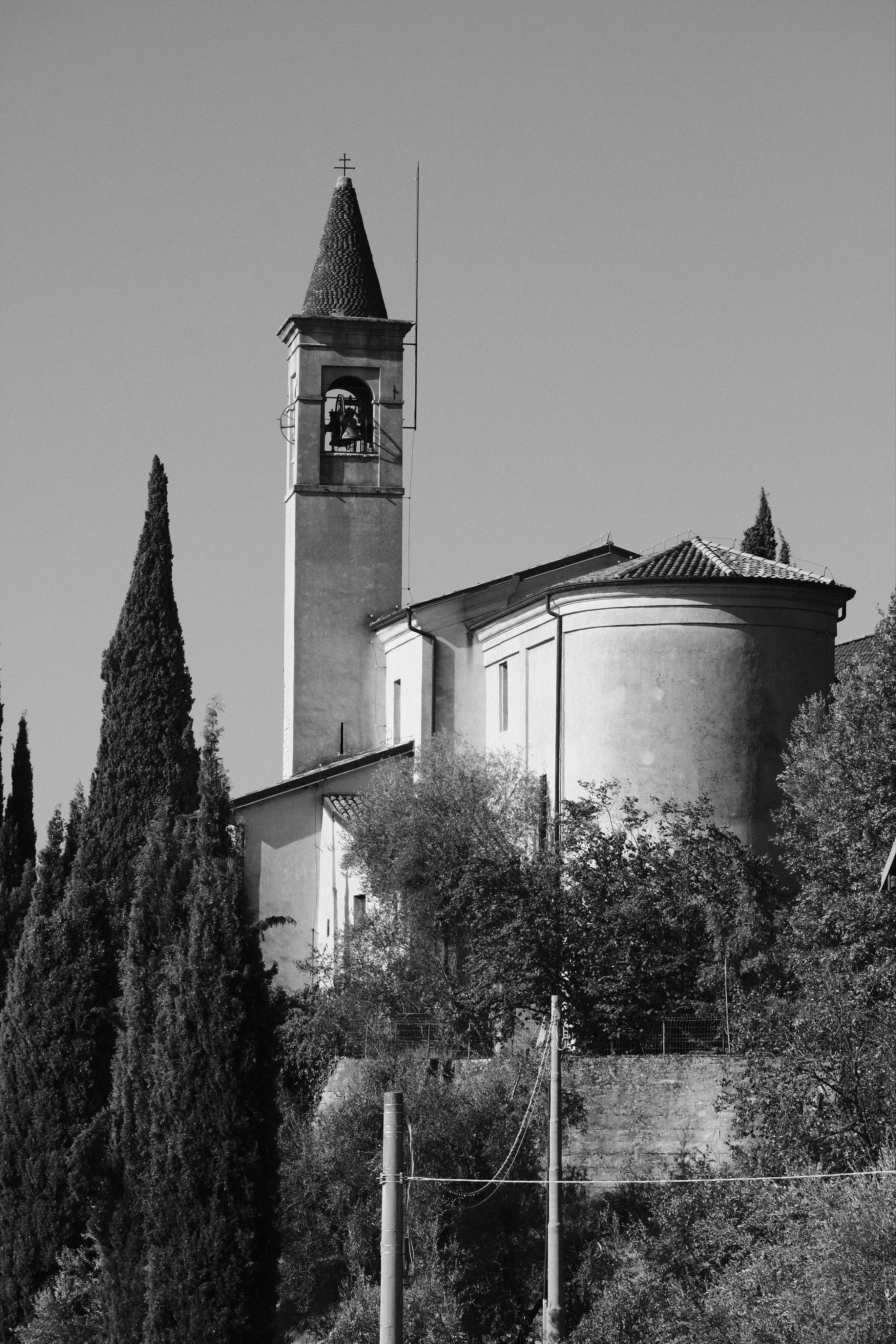 Black-and-white photo of a smaller church with a tall bell tower topped by a tiny cross, a bell visible in the arched opening, and a rounded apse with a tiled roof. The church is surrounded by cypress trees and other plants, under a clear sky.