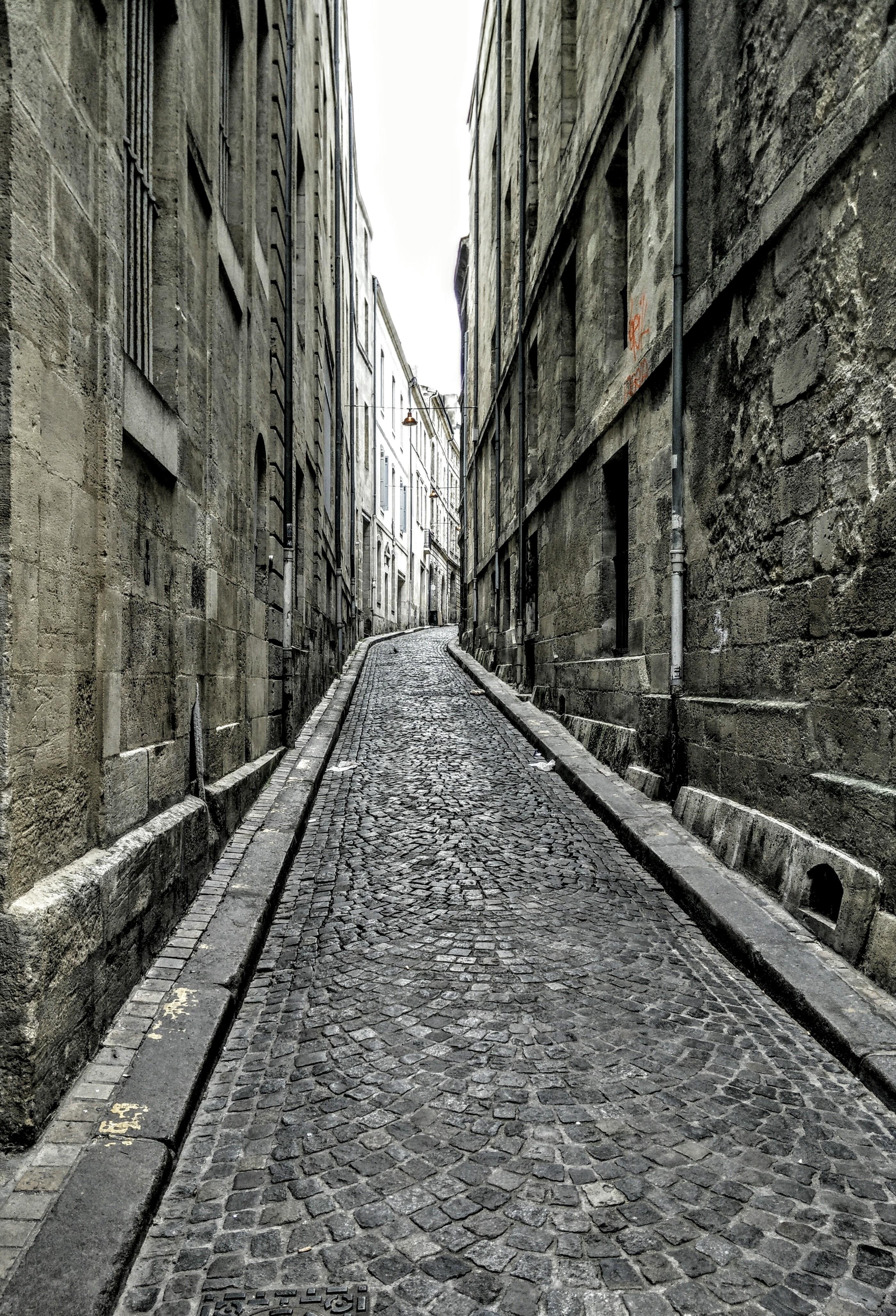 This image shows a narrow cobblestone street flanked by tall, weathered buildings. The street appears to be in an older part of a European city, characterized by stone architecture and a slightly worn, historic look. The path tapers off into the distance, slightly curving, and lined with stone gutters on either side.