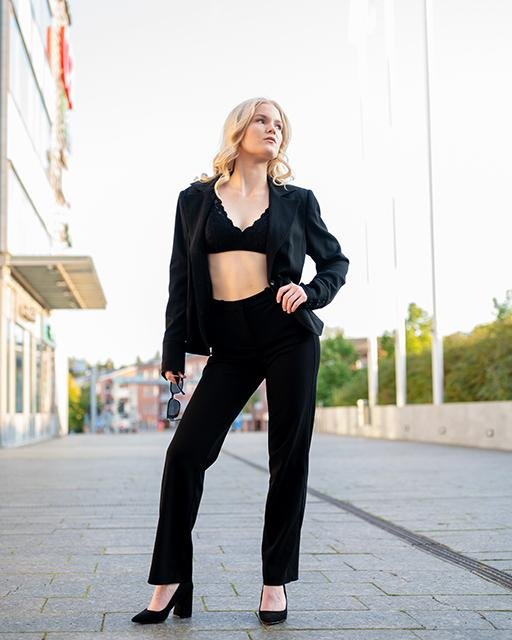 Blonde woman in a black outfit, including a cropped top and blazer, poses confidently on a city street. She holds sunglasses in her right hand and wears high heels. Modern buildings and greenery are visible in the background, with a clear sky overhead.