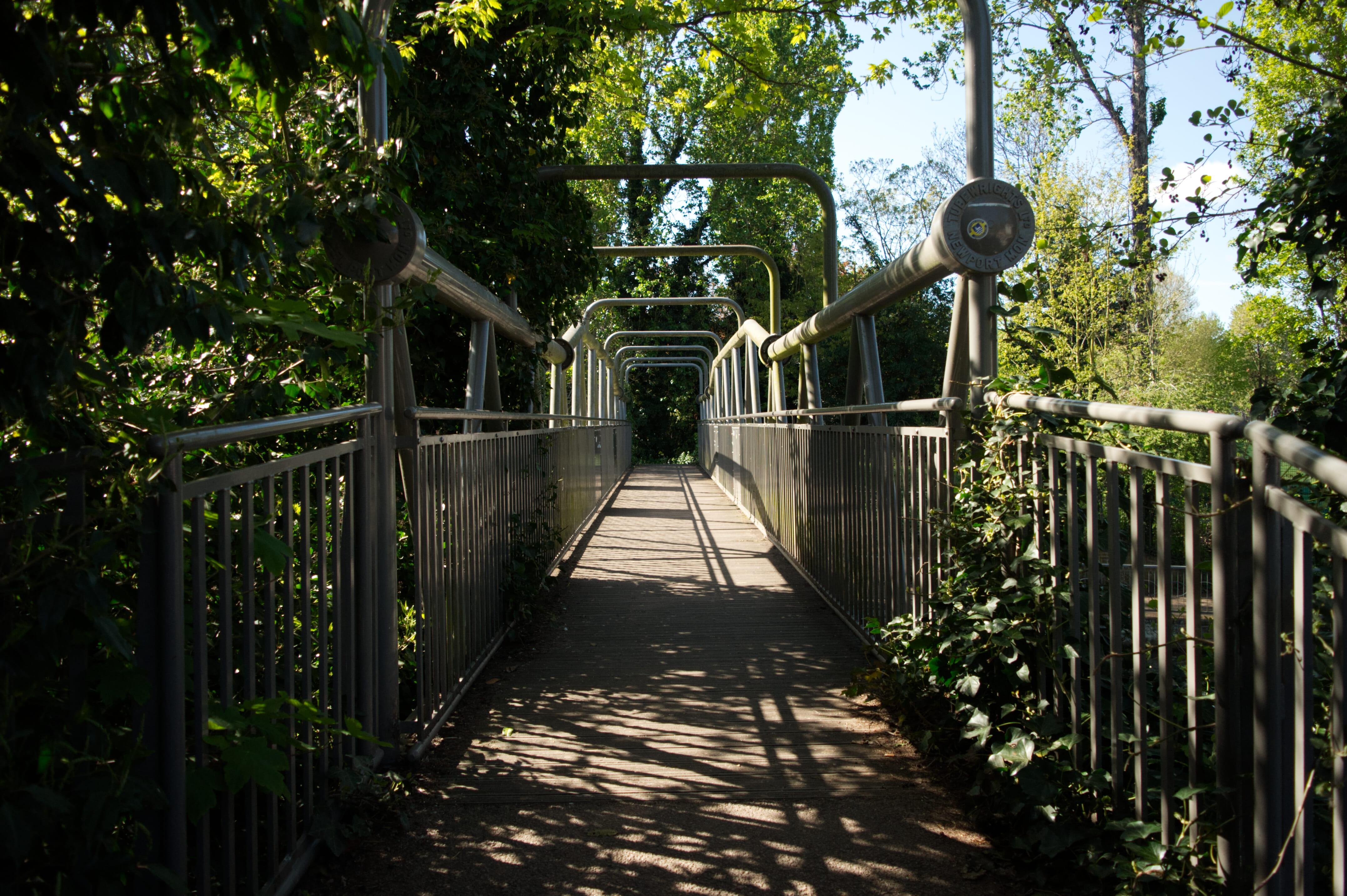 A view from the beginning of a small, metal-framed bridge hanging over a river. The metal is generally what appears to be an unpainted silver. There are barriers (also made of metal) that span the entire length of the bridge to prevent pedestrians from simply falling off. The frame itself creates a triangle pattern for the sides and is best seen on the other side of the barrier - however, it can be seen somewhat from this view. At the middle of some of the triangular frame sections are curved rectangular pieces which function somewhat as arches. Nearer the front, where one of the main tubular sections begins is a cap for one of the tubes that has "Tubewrights Ltd, Newport Mon" embossed into it, the words circling the cap. There is also an unknown yellow and blue sticker stuck to the cap, though it is illegible in the photo.

The bottom of the bridge does appear to be made of small, thin planks of wood.

There is foliage pushing its way through the right-hand barrier nearest the camera and it is slowly stating to push through on the left-hand side as well. There are trees everywhere, though the actual path the bridge creates is clear.

The sun is shining quite brightly here, so the path is lit up quite brightly. You can see the pattern of the barriers (tall, thin, rectangular) as the bars create small patches of shade. The trees also block out some of the sun nearest the camera, creating patches of shade and also showing some shadows of leaves.