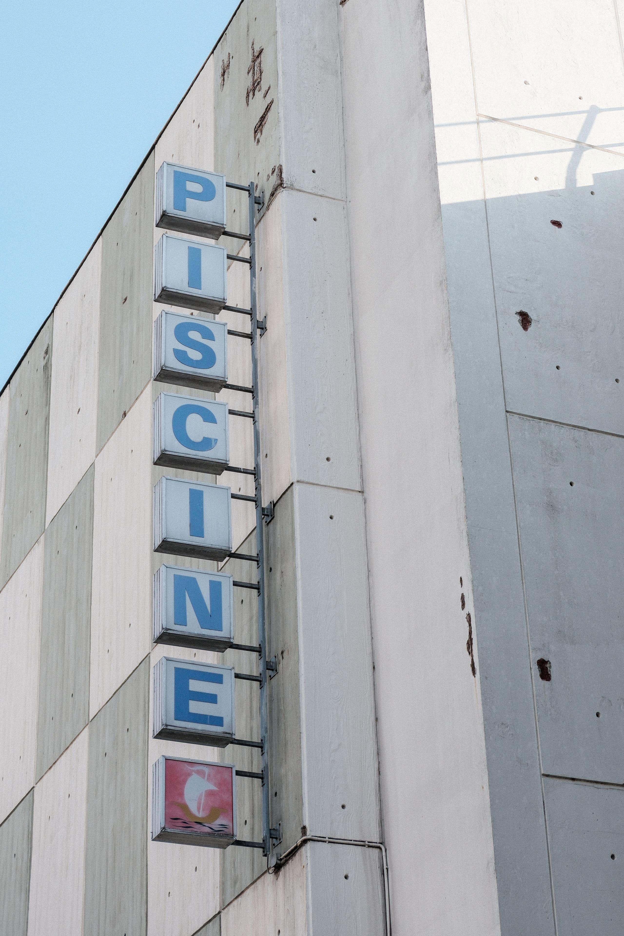 Photo verticale. L’enseigne d’une piscine qui écrit le mot piscine, 1 carré par lettre bleu sur fond bleu. Un dernier carré ponctu le mot d’un emblème blanc sur fond rouge
