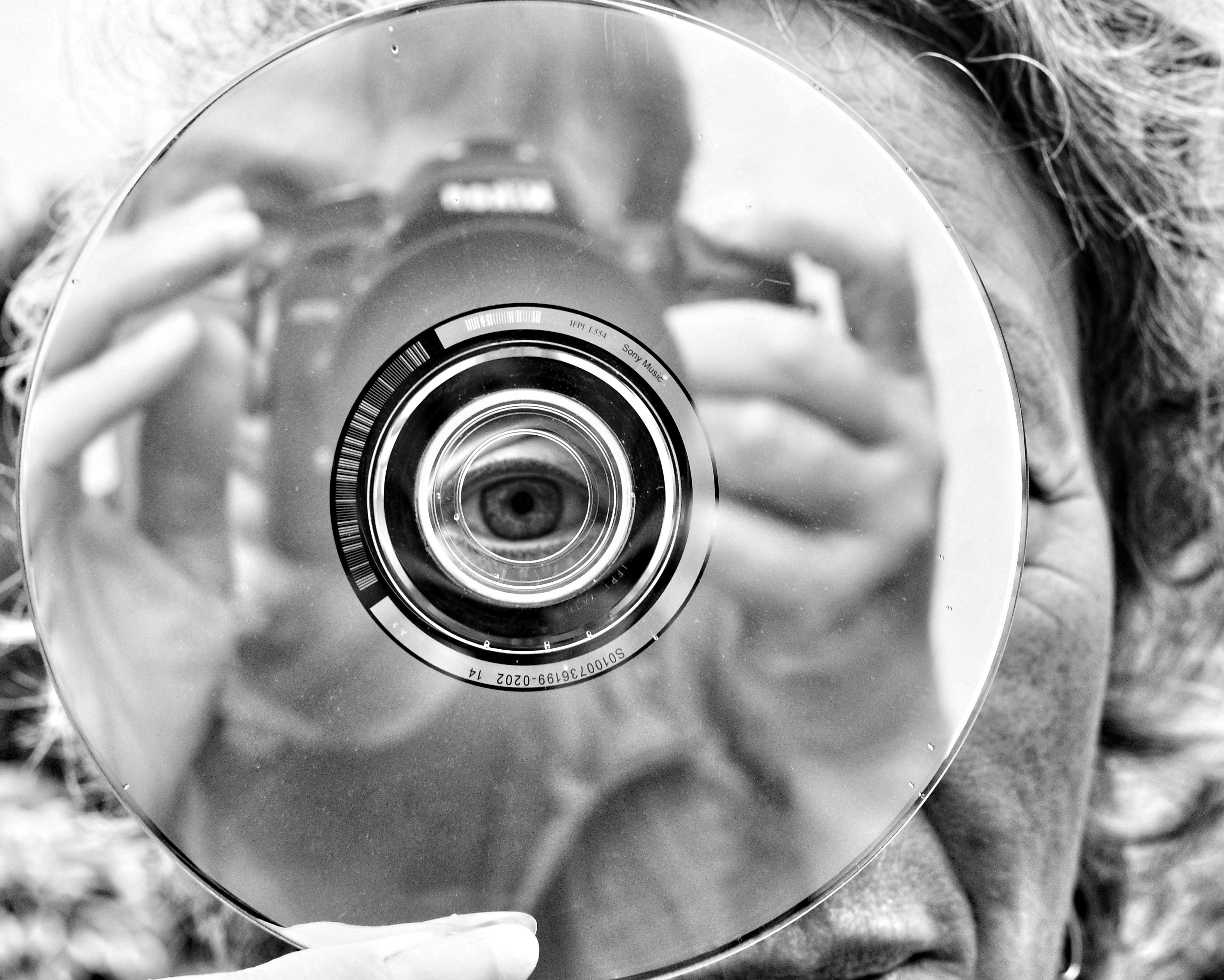 Black-and-white close-up of a person holding a camera, reflected in a compact disc; the disc’s center hole aligns with one eye, and the camera lens appears centered within the disc.###
Schwarz-Weiß-Nahaufnahme einer Person, die eine Kamera hält, die sich in einer CD spiegelt; das Mittelloch der CD ist auf ein Auge ausgerichtet, und die Kameralinse erscheint mittig innerhalb der CD.