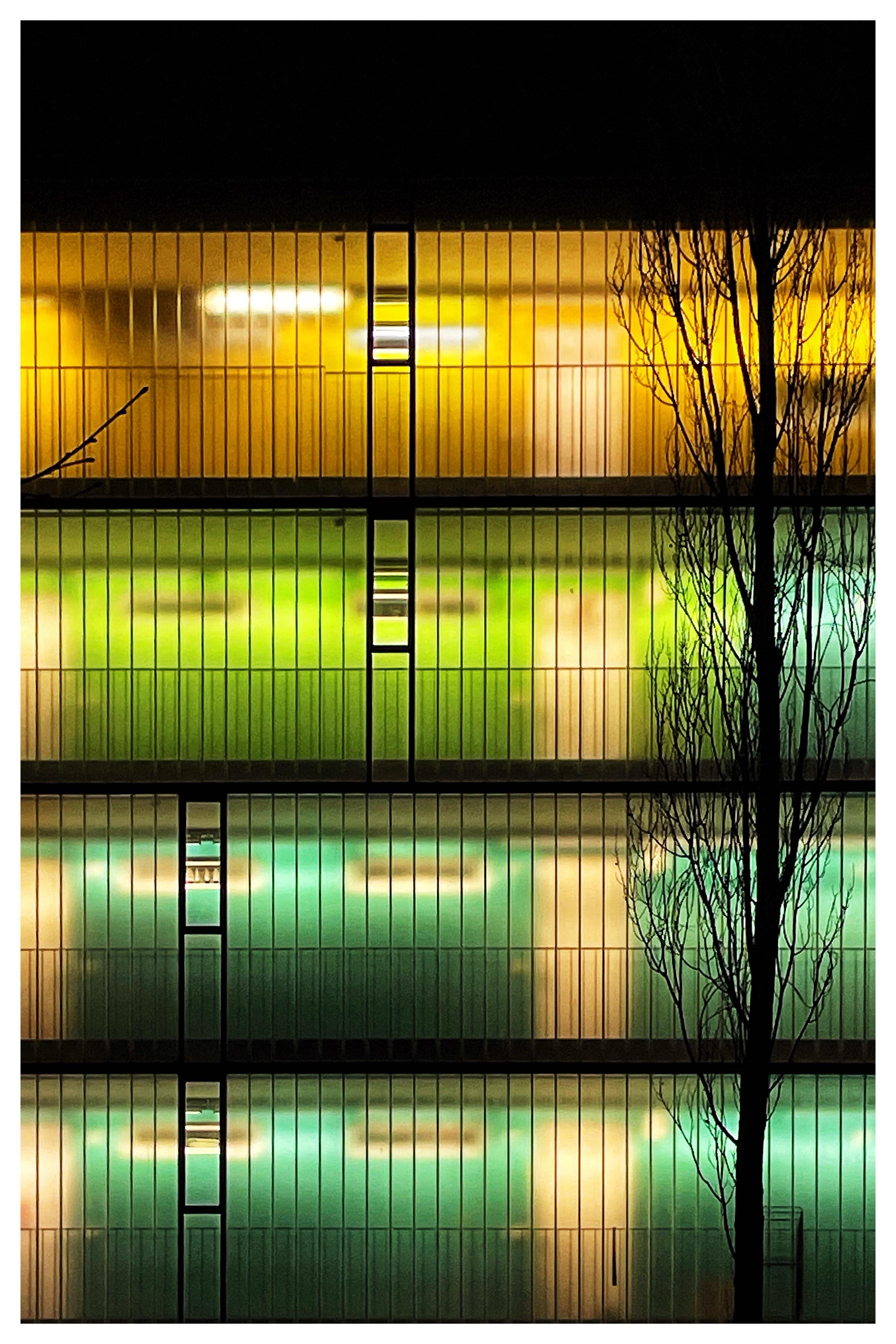 Multi-level building facade at night with vertical railings and glowing interior lights; top floor lit yellow, lower floors lit green and teal, with a leafless tree silhouette along the right edge and a narrow vertical column of windows near the center.