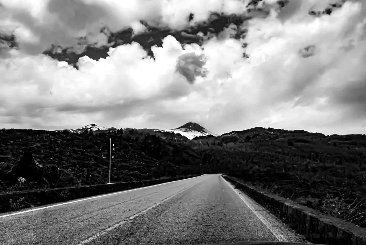 Black-and-white view from the center of an empty paved road stretching into the distance between low concrete barriers, leading toward dark forested hills and a snow-dusted mountain peak beneath large, layered clouds.