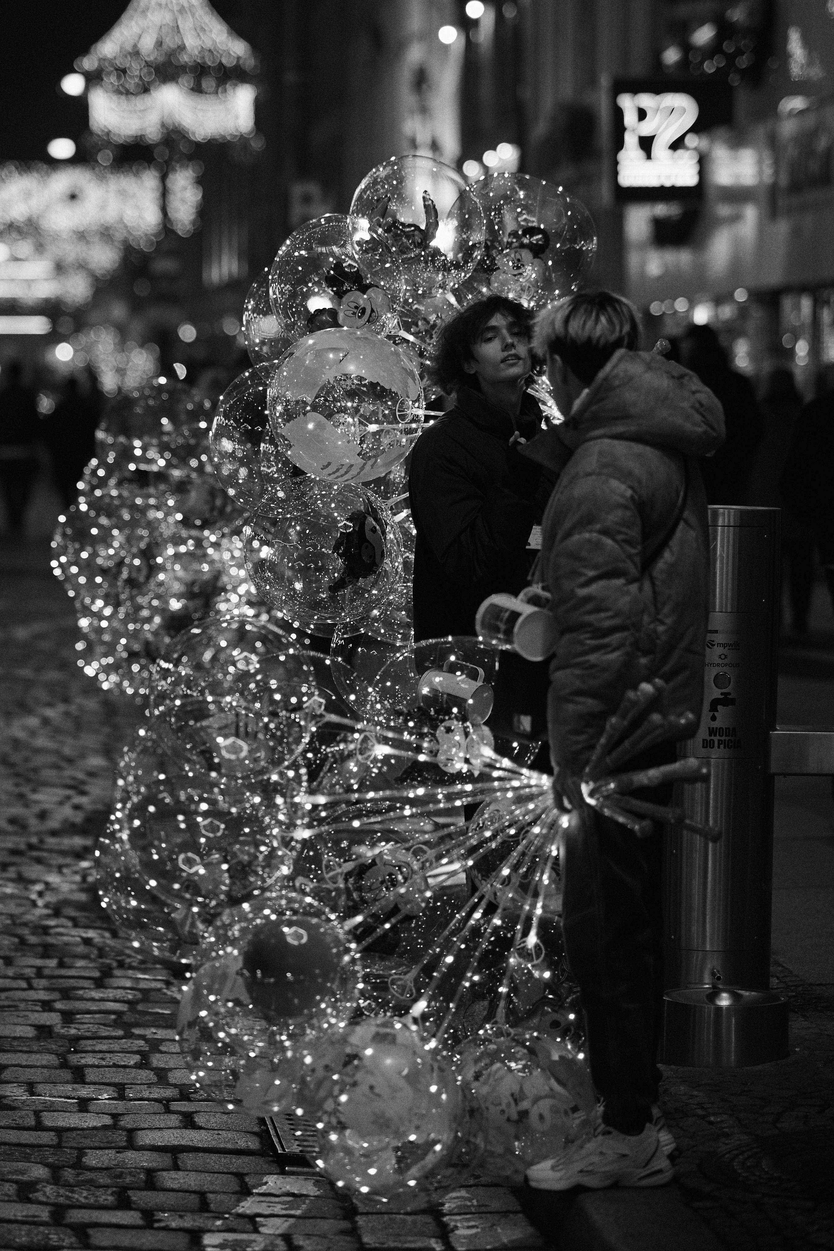 The black and white image shows a street scene with two people talking. One of them is holding a bunch of transparent balloons illuminated with small lights. The background has decorative lights and some out-of-focus signage, suggesting a festive atmosphere or a nighttime city setting. The street is cobblestone, and the overall scene has a lively, bustling feel.