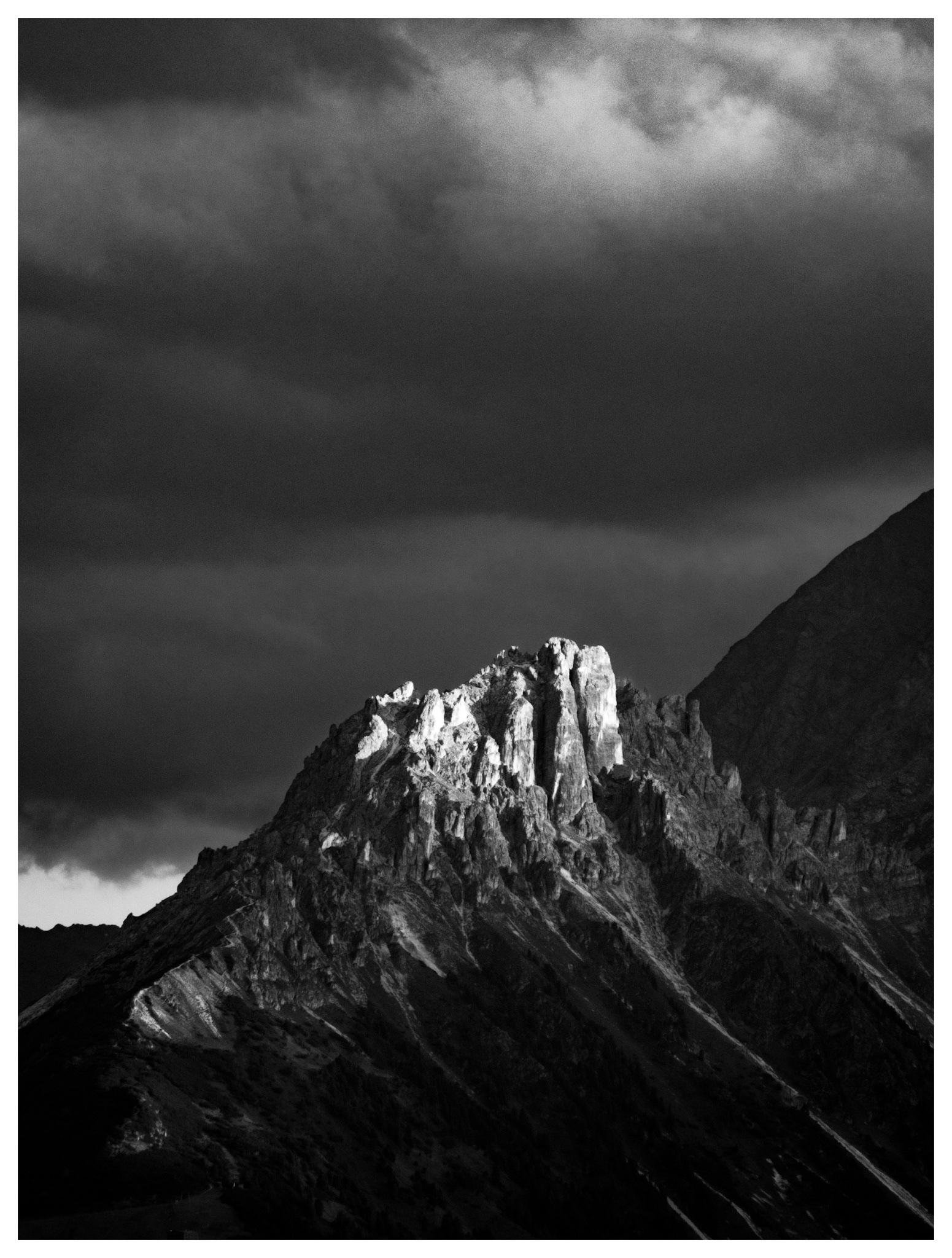 Black-and-white landscape photo of a jagged mountain peak with sunlit rock faces, set beneath heavy layered clouds; darker ridges and slopes fill the lower frame.