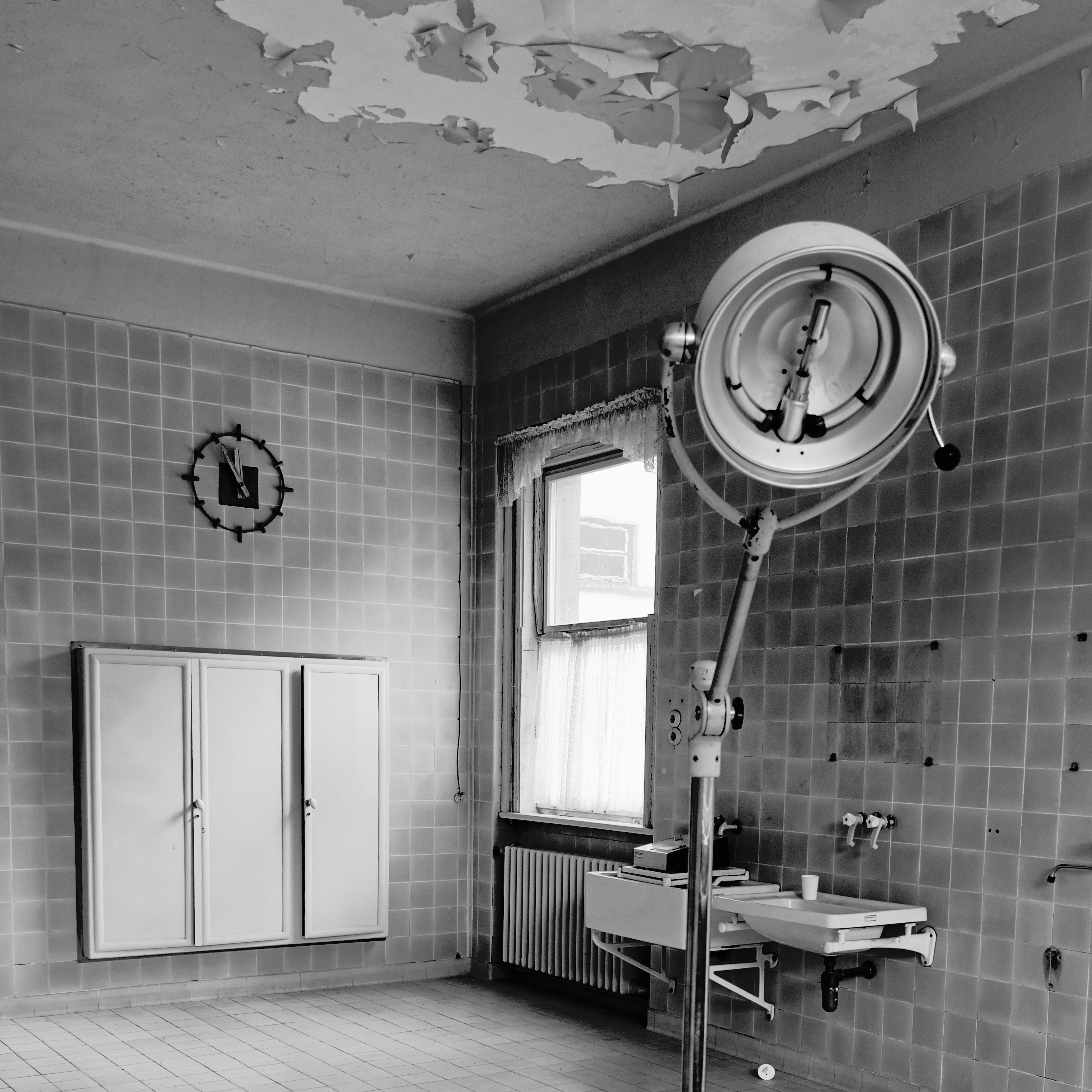 Old hospital room with peeling paint on the ceiling, tiled walls, and a large surgical lamp. A clock on the wall reads 1:00. A cabinet with closed doors is mounted on the wall, and a window with sheer curtains allows light into the room. A sink is visible to the right, beneath the window, with a radiator below it. 
### 
Altes Krankenhauszimmer mit abblätternder Farbe an der Decke, gefliesten Wänden und einer großen OP-Lampe. Eine Uhr an der Wand zeigt 1:00 Uhr an. An der Wand ist ein Schrank mit geschlossenen Türen montiert, ein Fenster mit durchsichtigen Vorhängen lässt Licht in den Raum. Rechts unter dem Fenster ist ein Waschbecken zu sehen, darunter befindet sich ein Heizkörper.