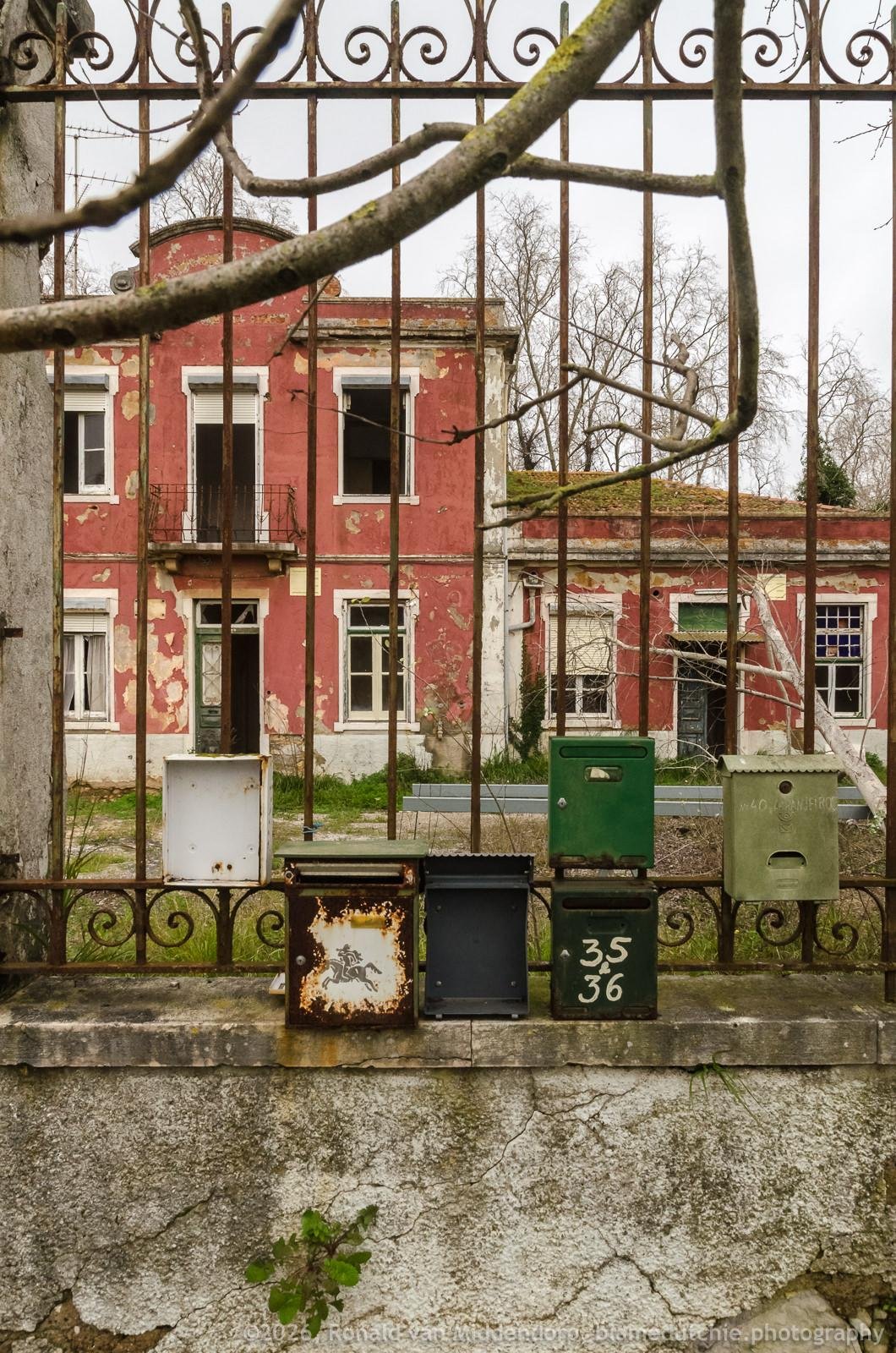 Six mailboxes mounted on an iron fence with curled detailing in the foreground and bare tree branches crossing the top, in front of a weathered red two-story building with peeling paint and several broken or open windows and doors.