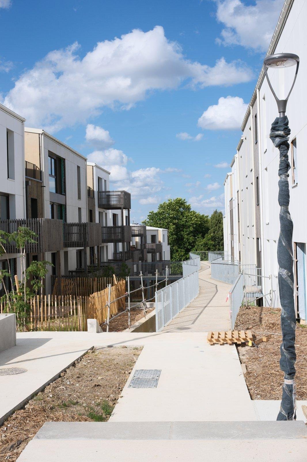 Photo format portrait montrant une résidence d’immeubles de 3 étages en cours de finition, on voit un lampadaire au premier plan, et une passerelle qui part au loin