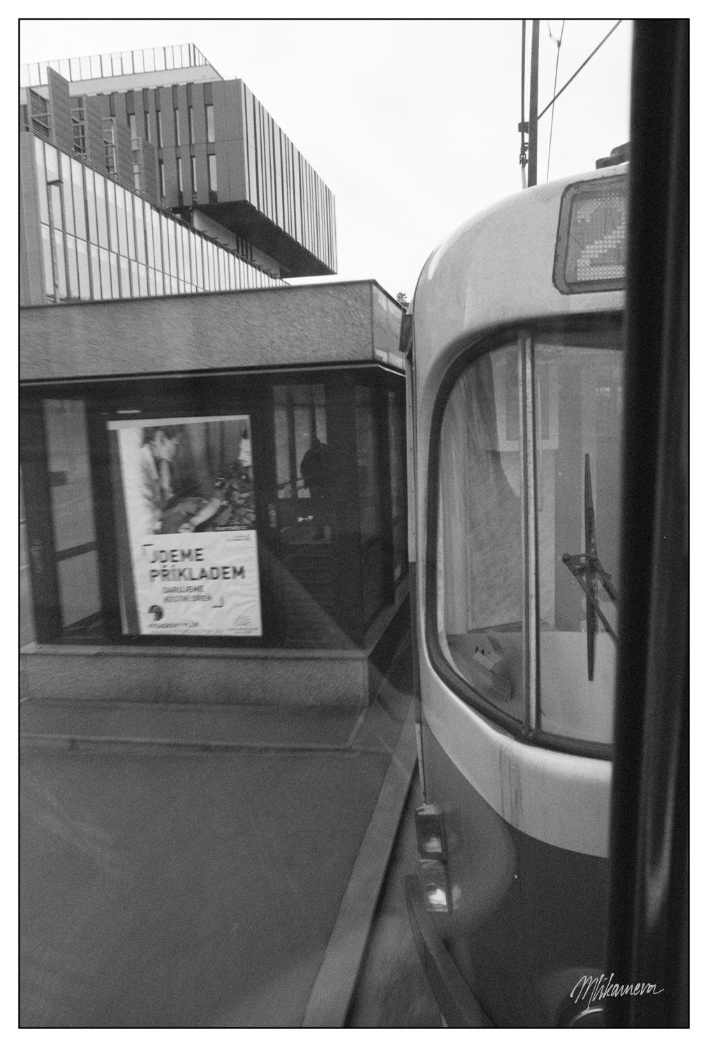 Black and white image of a tram's front right corner in the foreground. A modern, multi-story building with vertical and horizontal lines is in the background. A bus stop shelter with a poster, possibly displaying an advertisement featuring a person, is visible near the tram. Wires are stretching above the tram, and a sidewalk is visible beside the shelter.