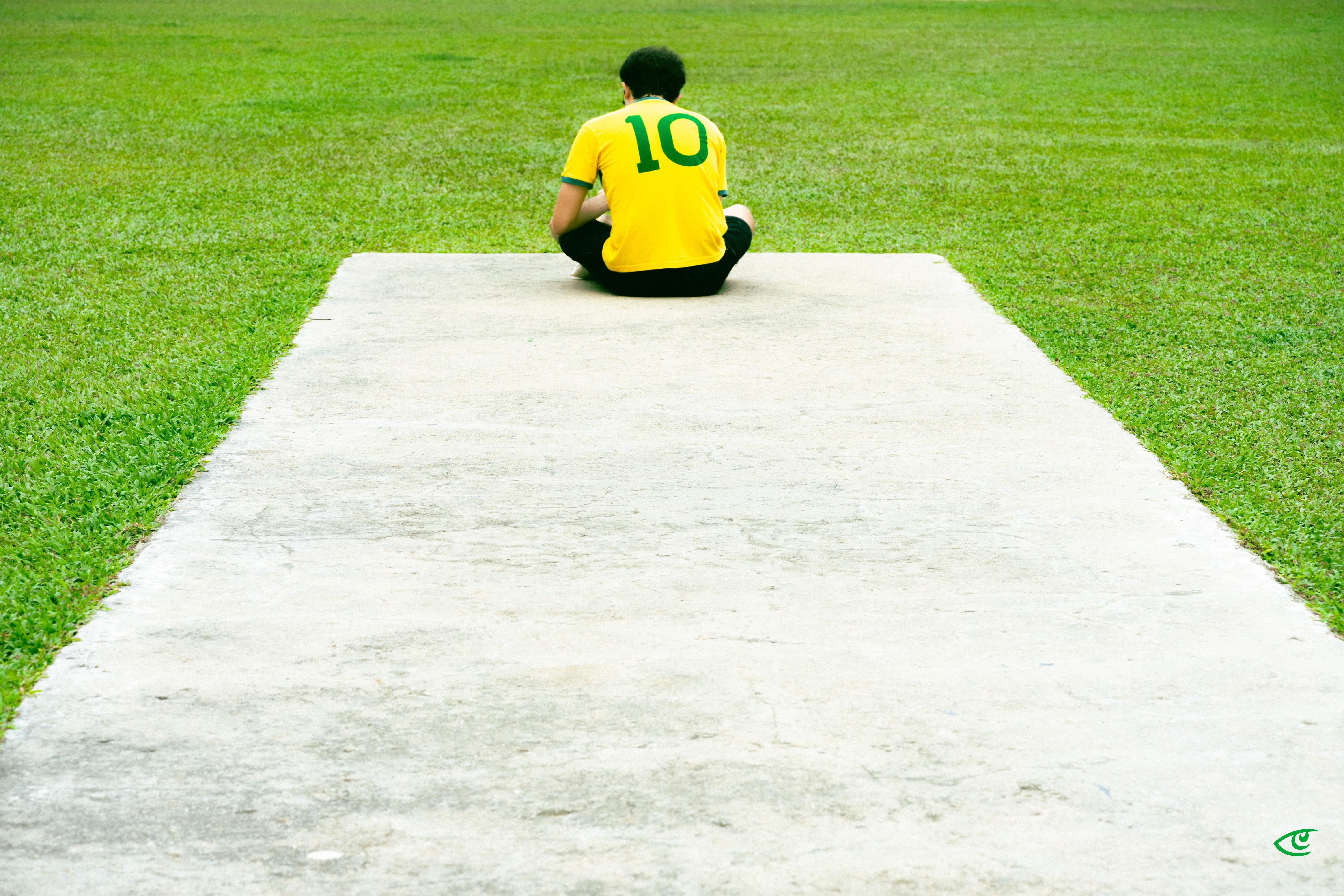Person sitting cross-legged at the far end of a wide concrete path, viewed from behind, wearing a yellow jersey with a green number 10 and black pants, surrounded by green grass.