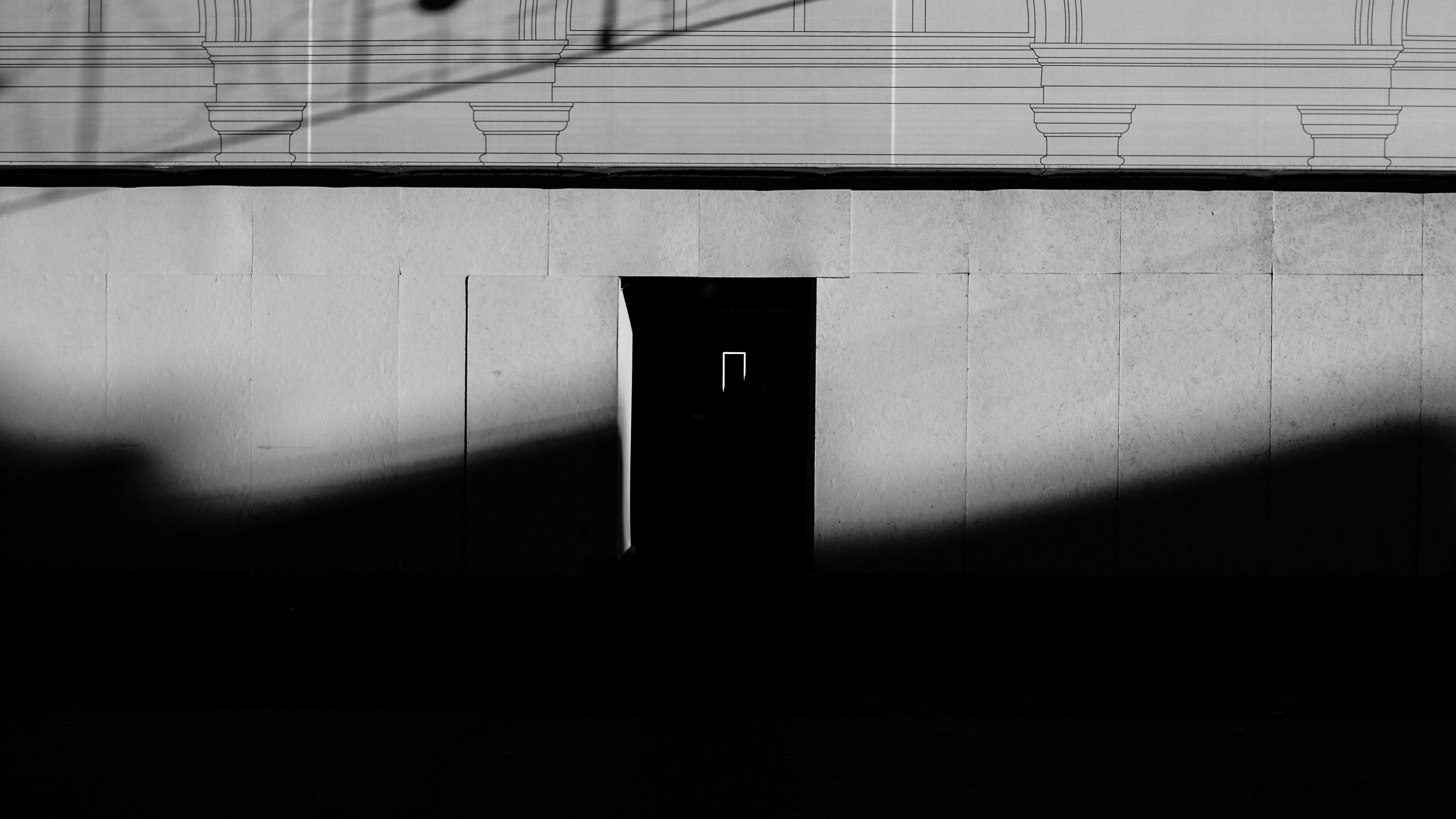 The image features a black and white photograph of a minimalist structure with a rectangular door or opening in the center. The light creates strong shadows and a high contrast pattern on the wall, emphasizing geometric shapes and lines.