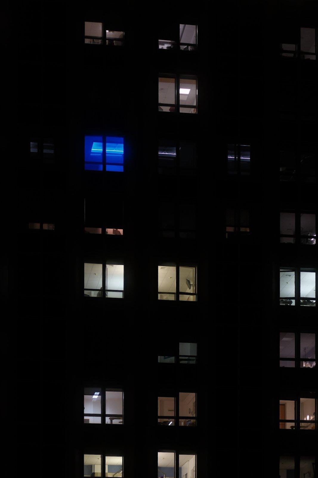 Photo verticale de nuit, façade de bâtiment avec plein de fenêtres allumées, une seule est en lumière bleu