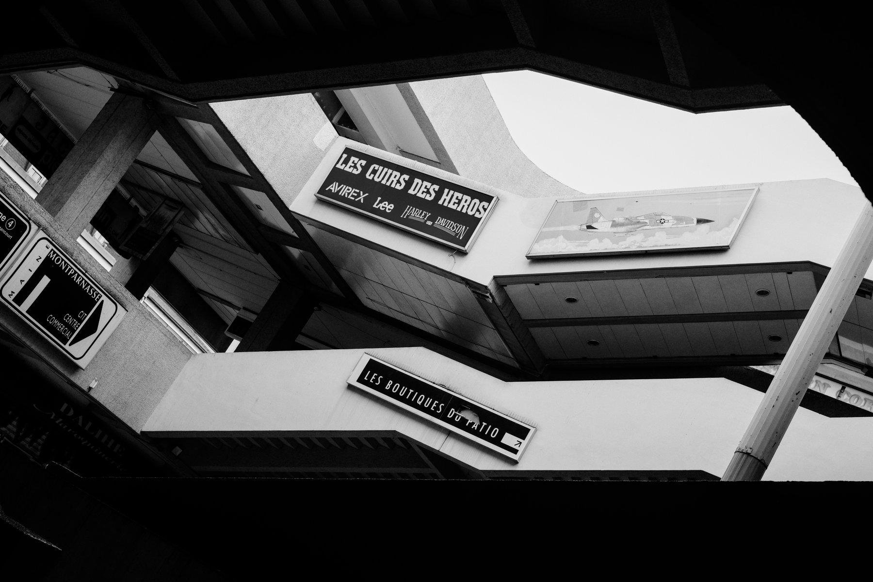 Photo horizontale en noir et blanc. Le haut et le bas sont totalement noir, et au centre, de la lumière et 2 étages en mezzanine, avec des enseignes sur les muret de sécurité, « Les cuirs des héros », « Les boutiques du patio » et une sans nom mais avec un avion de chasse de profil