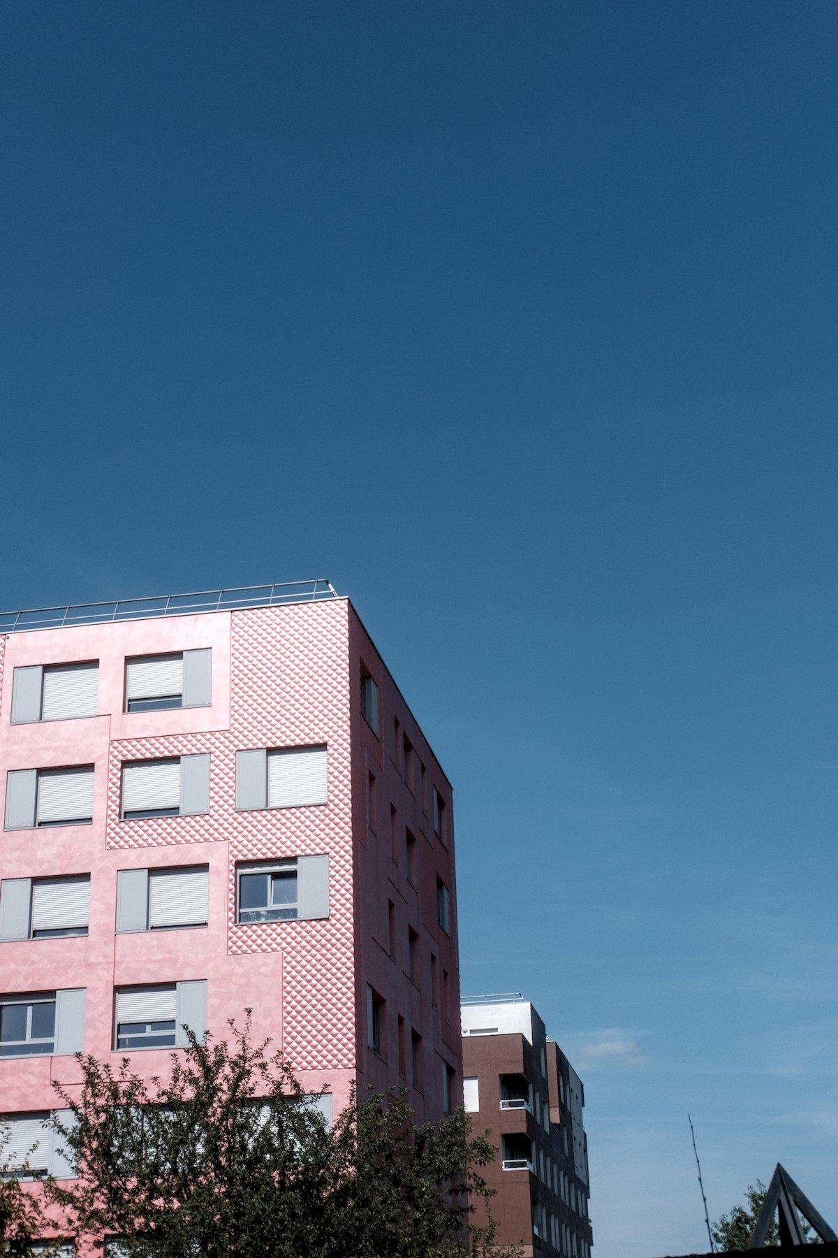 Photo verticale. Les 3/4 sont fait d’un dégradé de ciel bleu foncé en haut vers des nuages blanc en bas à droite. Le quart SW est un bout d’immeuble à la façade rouge