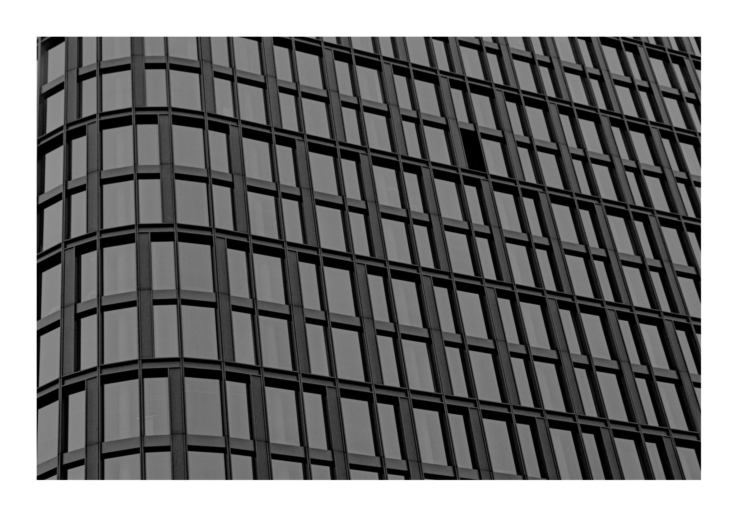 The image shows a black and white photograph of the facade of a modern building. The building has a grid-like pattern created by numerous windows and vertical as well as horizontal structural elements. The windows are arranged in a repetitive manner, contributing to a structured and geometric aesthetic.