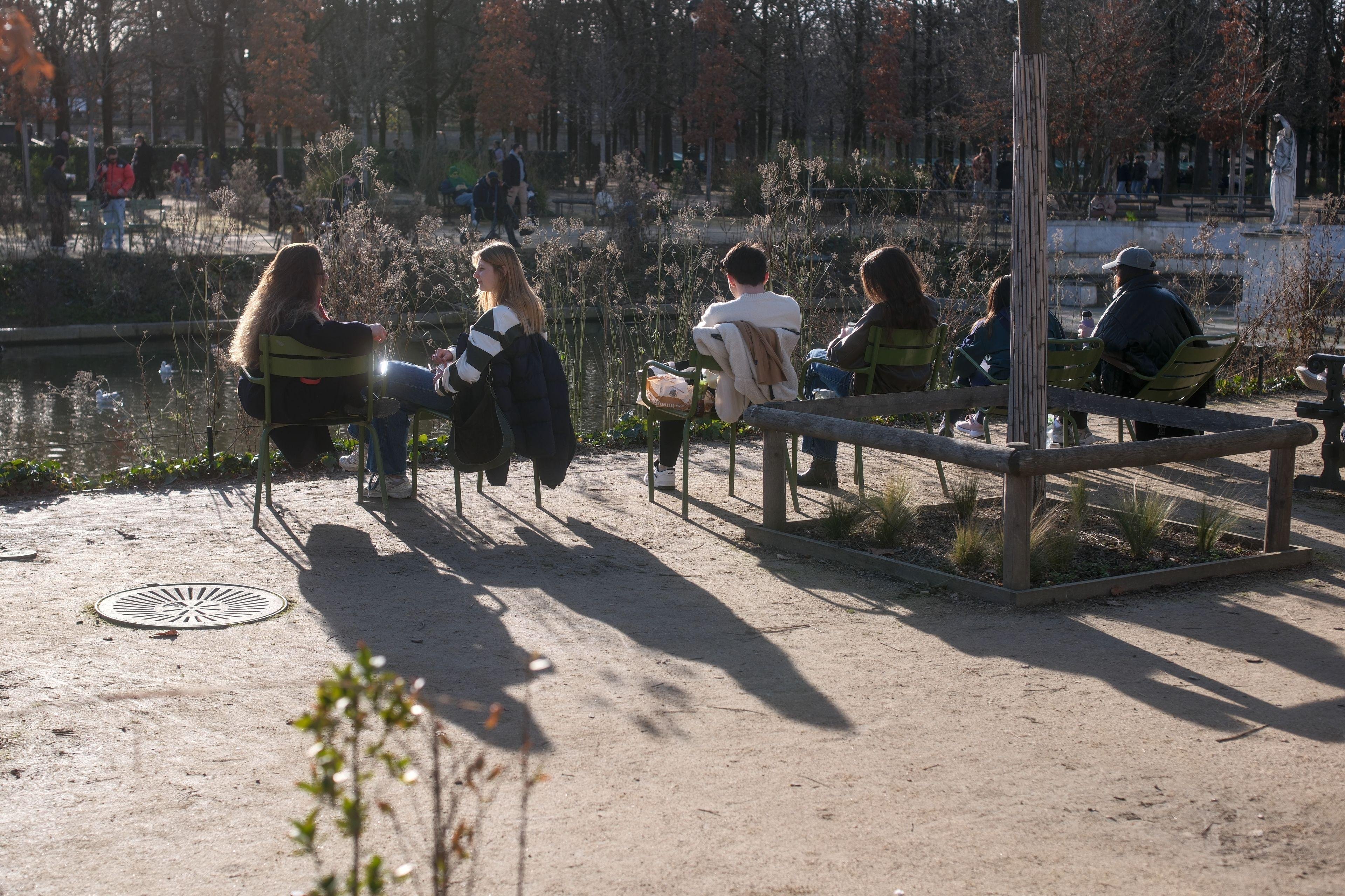 Photo horizontale. Des gens assis sur des chaises vertes en métal dans le parc du Luxembourg et plus particulièrement 2 femmes de profils en train de discuter, devant un plan d’eau, leurs longues ombres projetées sur le sol