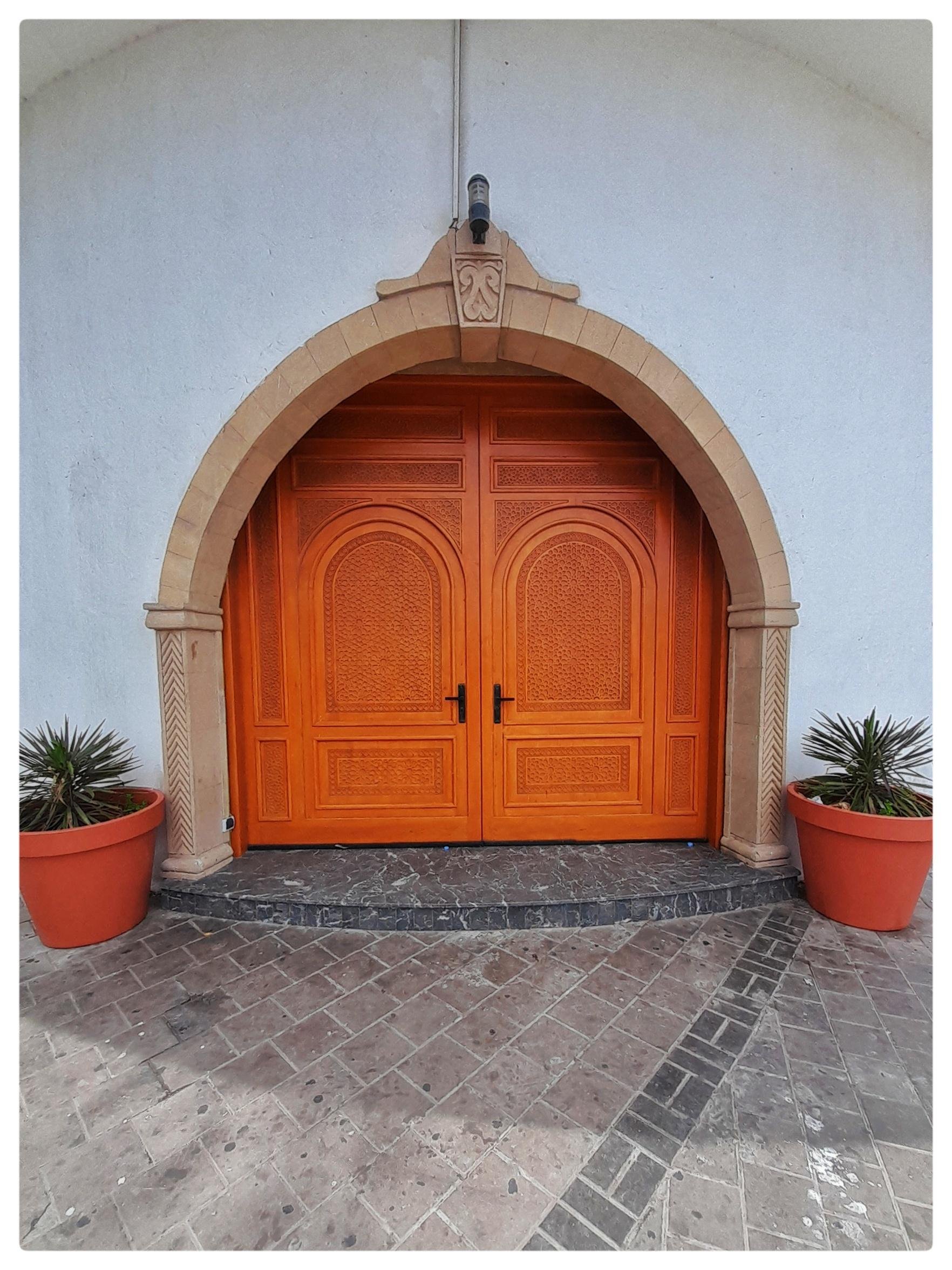 Arched stone doorway with carved details framing a pair of orange wooden double doors with black handles, set in a white wall; two large terracotta planters with spiky plants on either side and a tiled stone walkway in front.