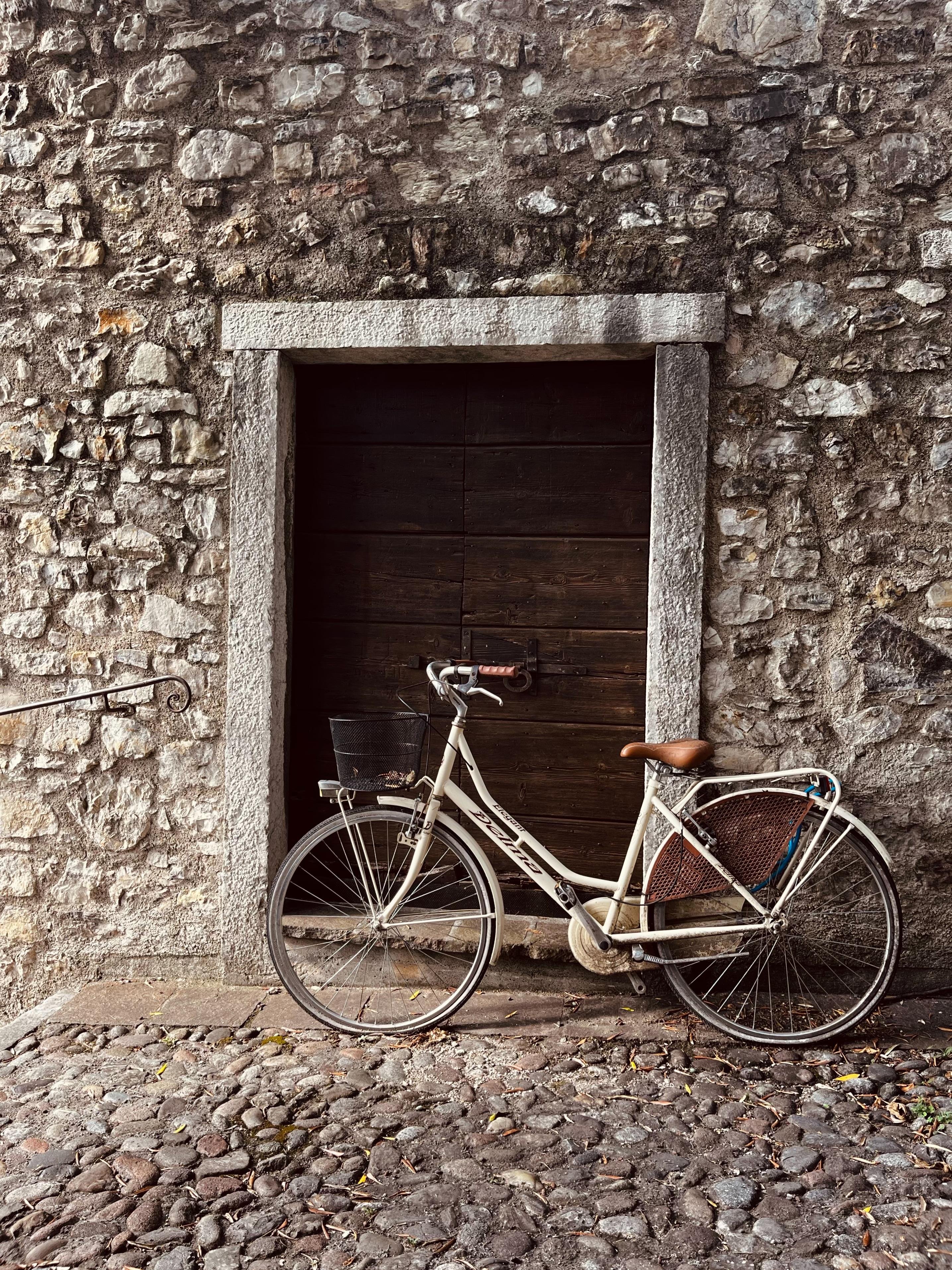 A beige, partly rusty bike leaning against a masonry stone wall with an ancient looking, heavy wooden door. The floor is covered in cobble stone of the Same colour as the wall.