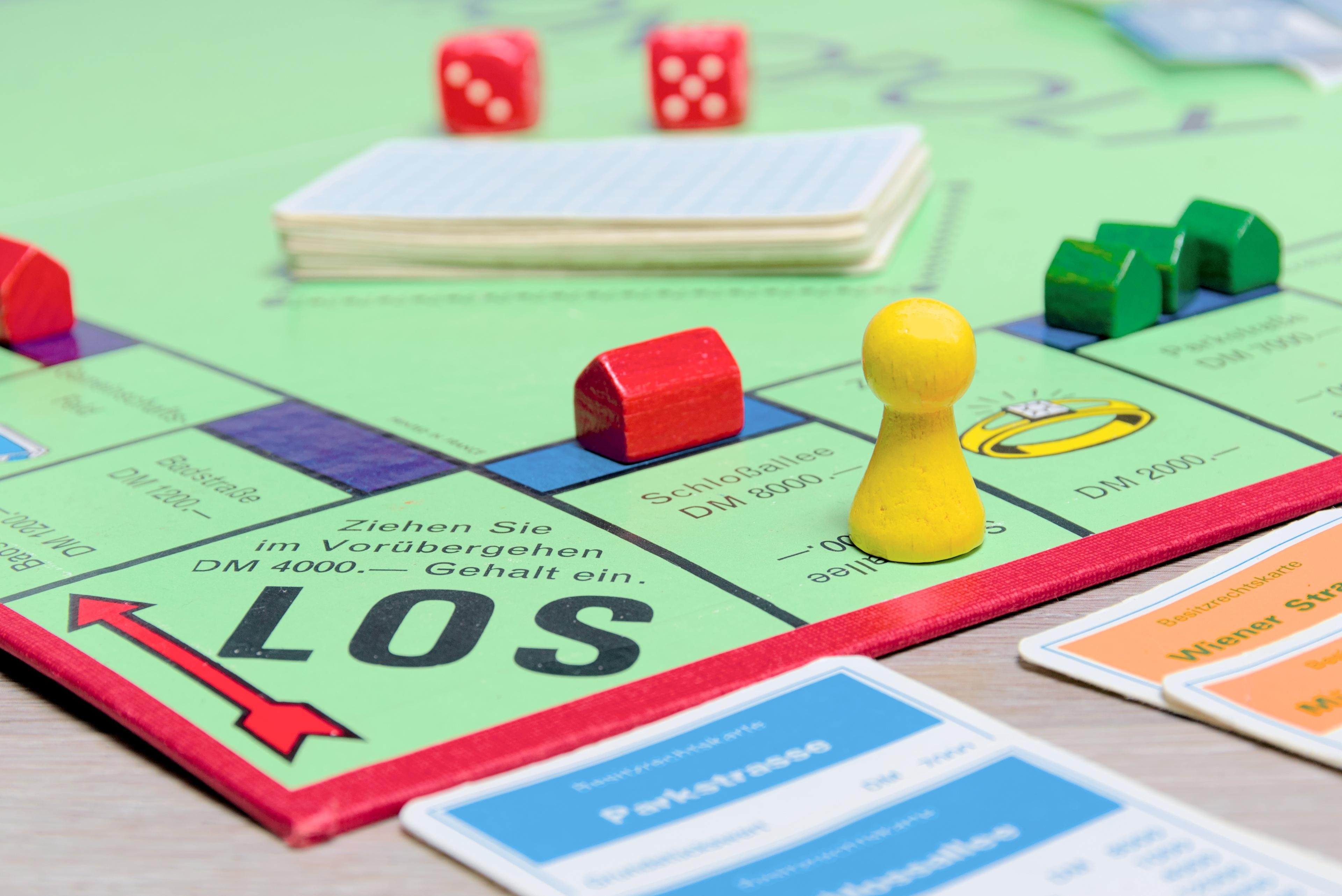 Monopoly board game setup with German text, featuring a yellow game piece, red dice, red house, and green hotels. Cards are stacked in the center; property cards labeled "Parkstraße" and "Wiener Straße" are visible in the foreground.
###
Monopoly-Brettspiel-Setup mit deutschem Text, bestehend aus einer gelben Spielfigur, roten Würfeln, einem roten Haus und grünen Hotels. In der Mitte sind Karten gestapelt; im Vordergrund sind Grundstückskarten mit der Aufschrift „Parkstraße“ und „Wiener Straße“ zu sehen.